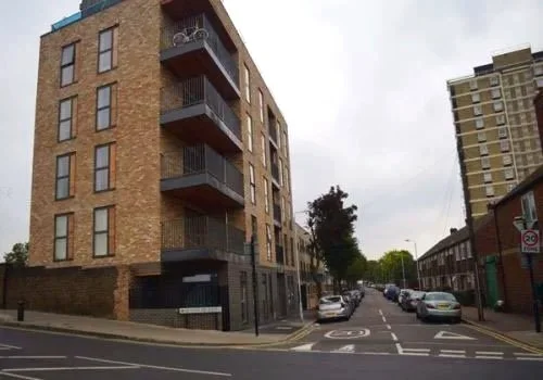 Urban street corner with multi-story brick apartment building, parked cars, and other buildings in the background.