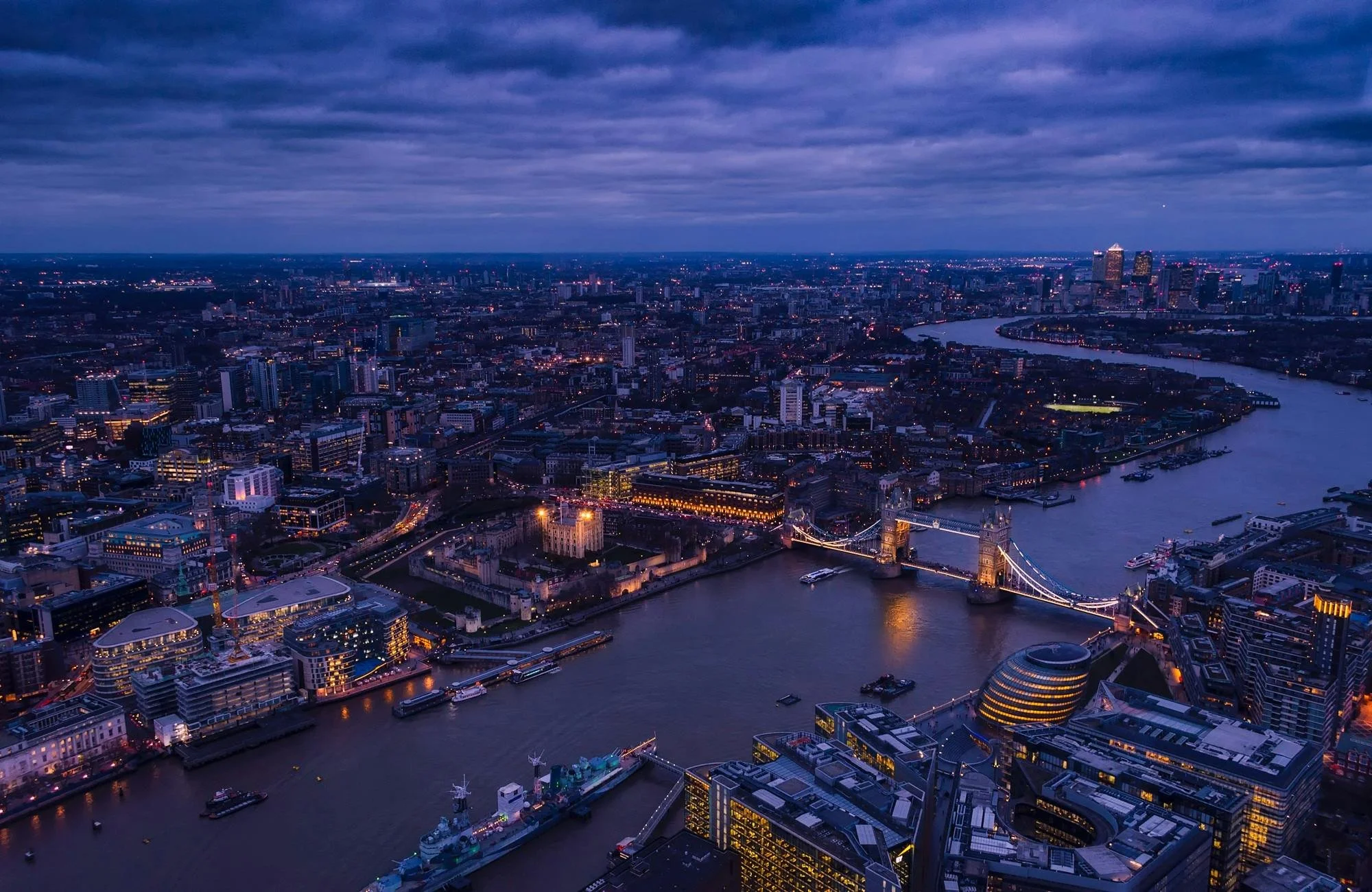 Aerial night view of London with Tower Bridge over the River Thames, illuminated cityscape, and cloudy sky.