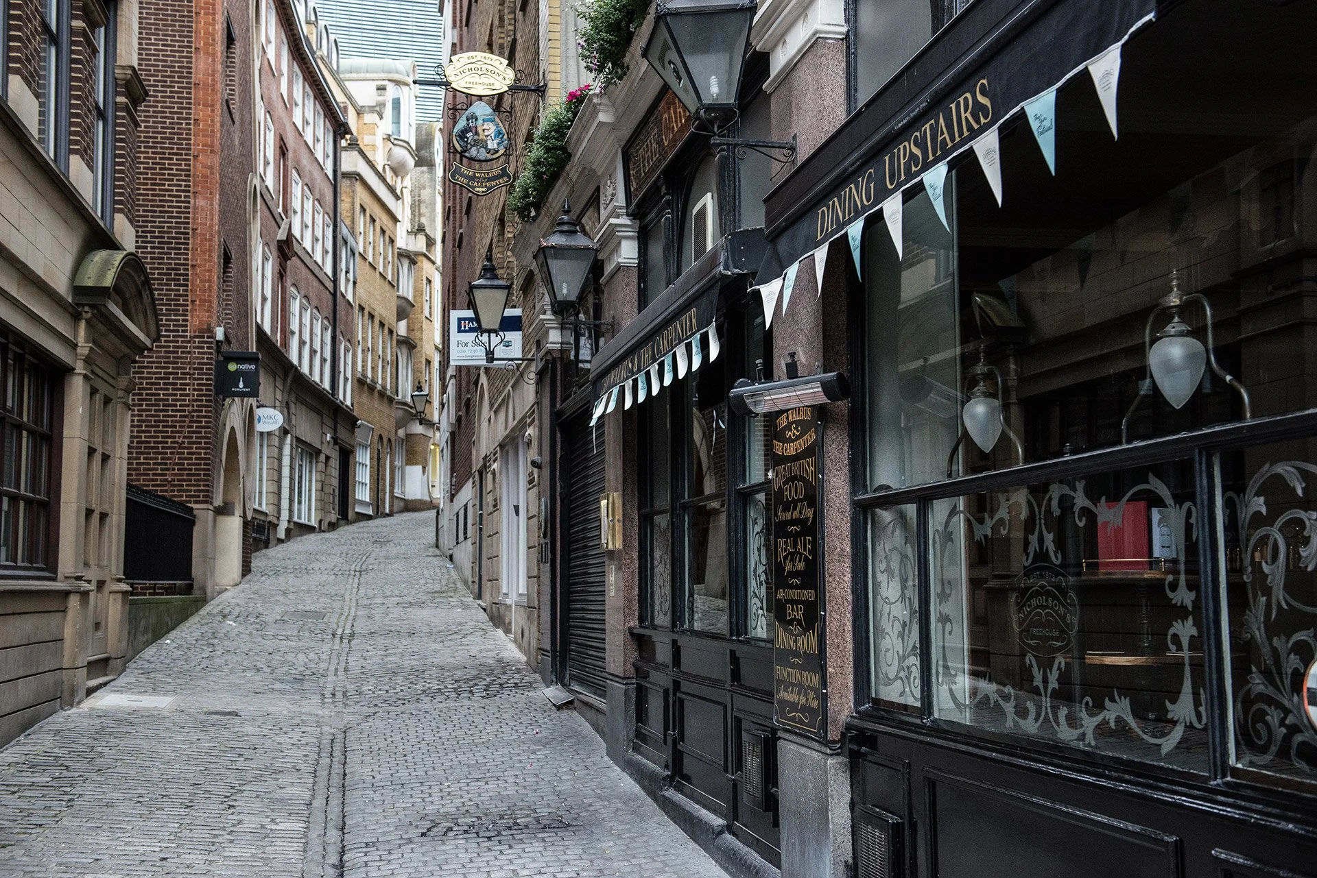 Empty cobblestone street alley with buildings on both sides, featuring a restaurant with hanging lamps and a black awning in the foreground, and multiple signs hanging from the buildings.
