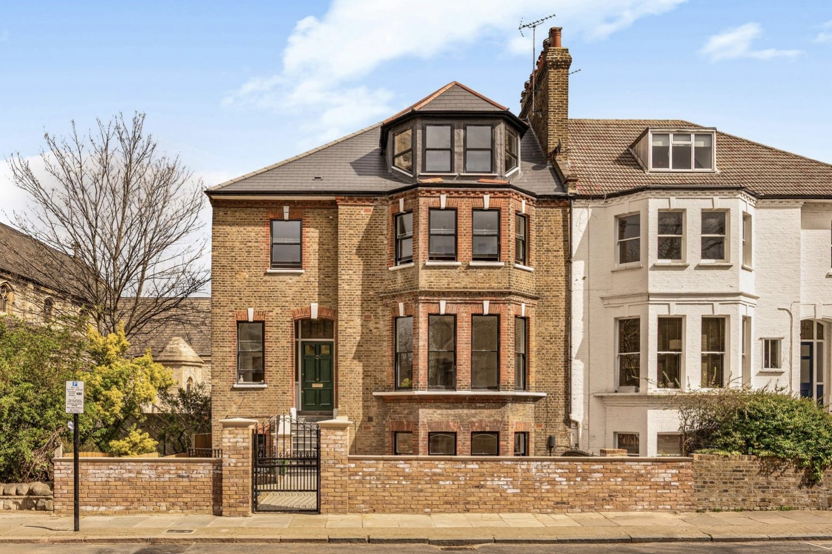 A three-story brick house with a green front door, black-framed windows, and a small front yard with a brick wall and gate. Adjacent white house with similar architecture is visible on the right, with leafless and green shrubs and trees around.