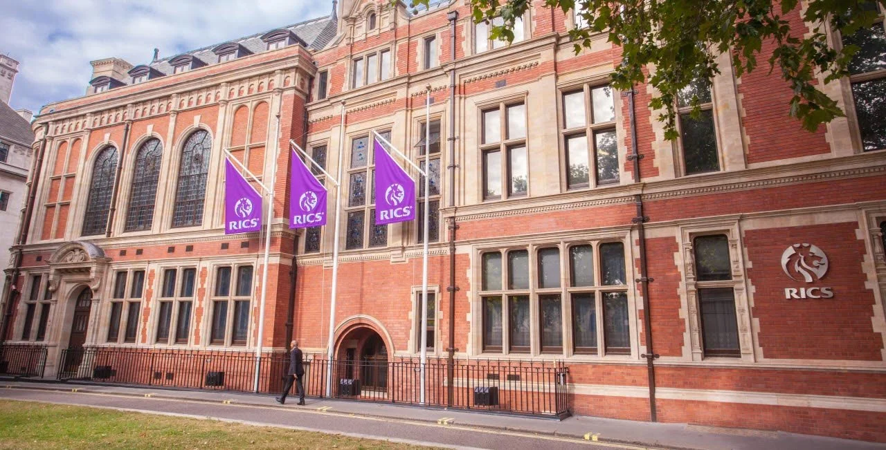 Red brick building with tall arched windows, purple flags with RICS logo, and a man walking on the sidewalk.