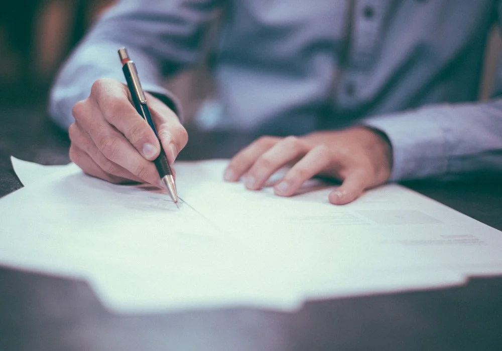 A person in a blue button-up shirt writing or signing on a document with a black and gold pen.