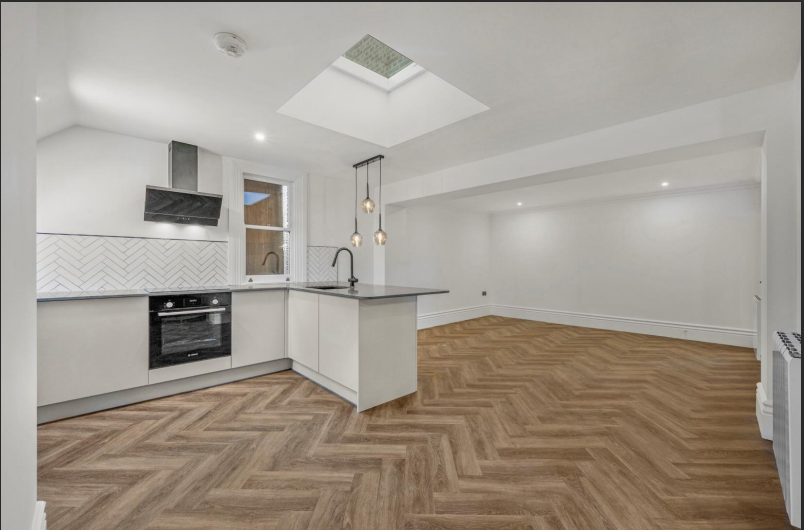 Modern kitchen and living space with white walls, wooden herringbone floor, skylight, and pendant lights.
