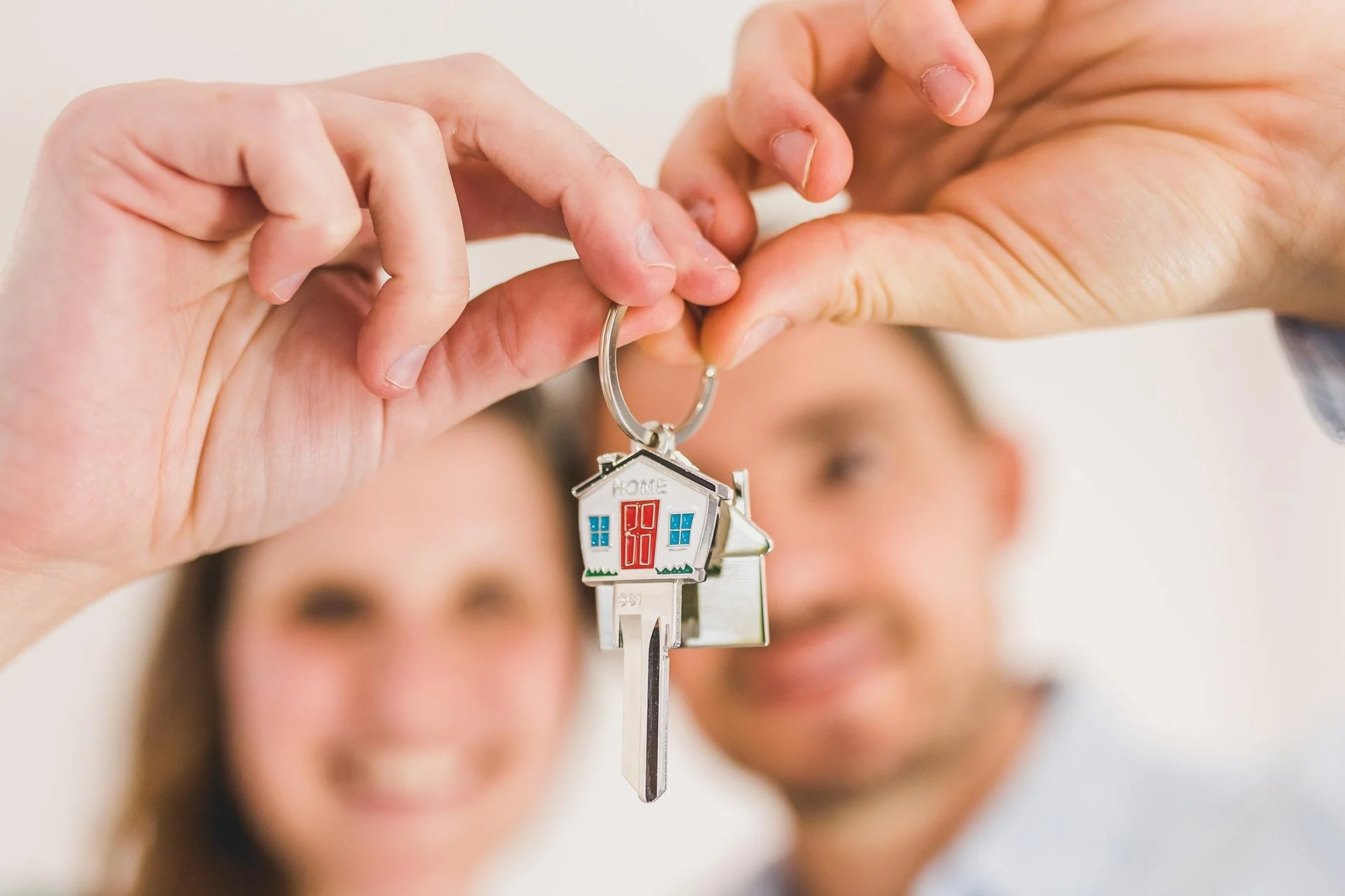 A close-up of a hand holding a house-shaped keychain with a key hanging from it, with smiling people blurred in the background.