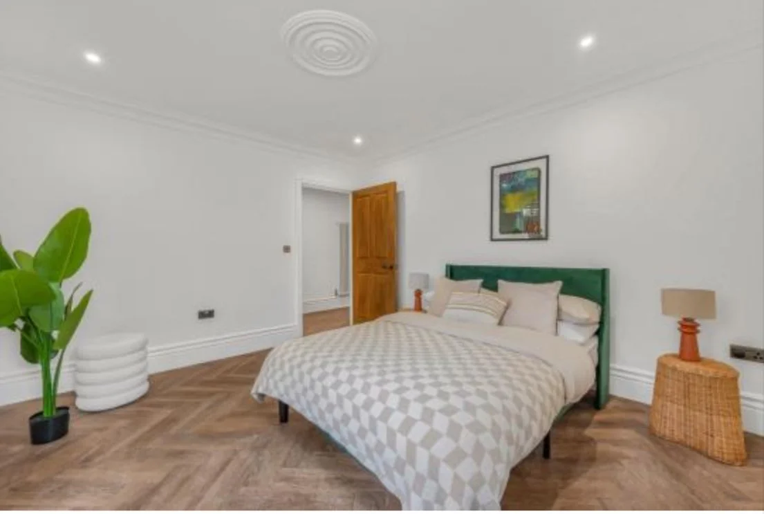 A bedroom with white walls and wooden flooring. A bed with beige bedding and pillows, a green headboard, and a framed artwork above. On each side of the bed are nightstands with lamps. There is a large green potted plant on the left, a small white st