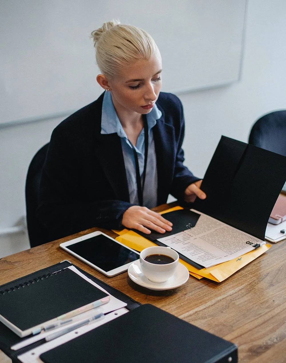 A young woman with blonde hair tied back, wearing a dark blazer and light blue shirt, sitting at a wooden desk reviewing documents with a focused expression. There is a white coffee cup, a tablet, and office supplies on the desk.
