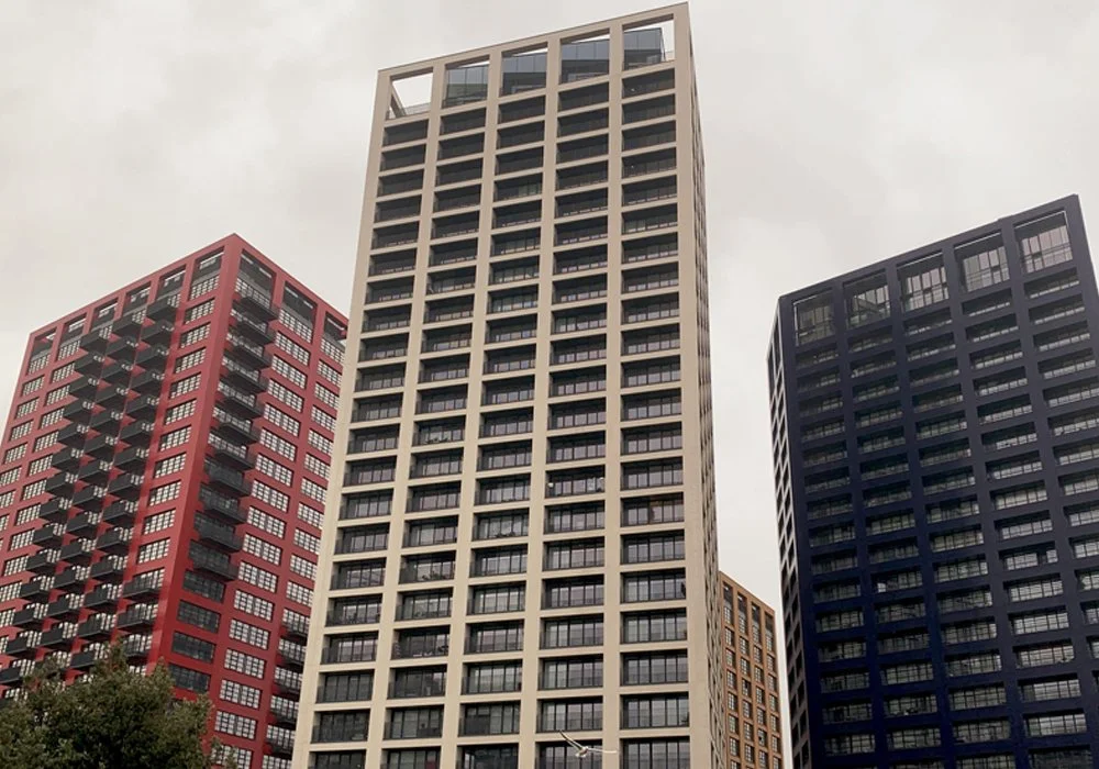 Three tall apartment or office buildings with grid-like window patterns, one beige, one red, and one dark blue, under a cloudy sky.
