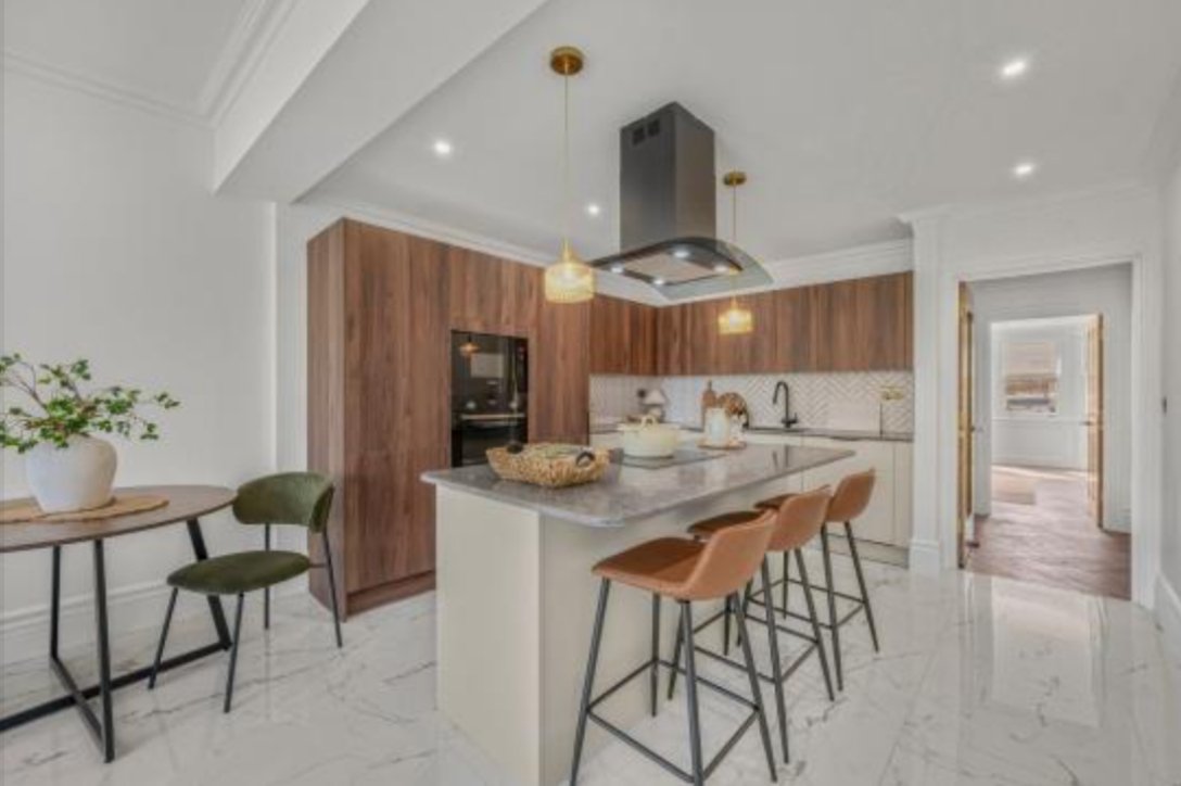 Modern kitchen with wooden cabinets, marble island with four brown bar stools, white walls, and a small dining table with green chairs and a potted plant.