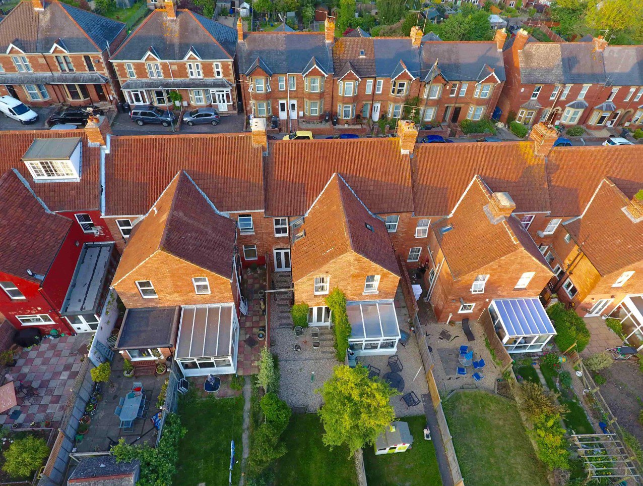 Aerial view of a row of terraced houses with brick facades and red tiled roofs, showing backyards, patios, and parked cars.
