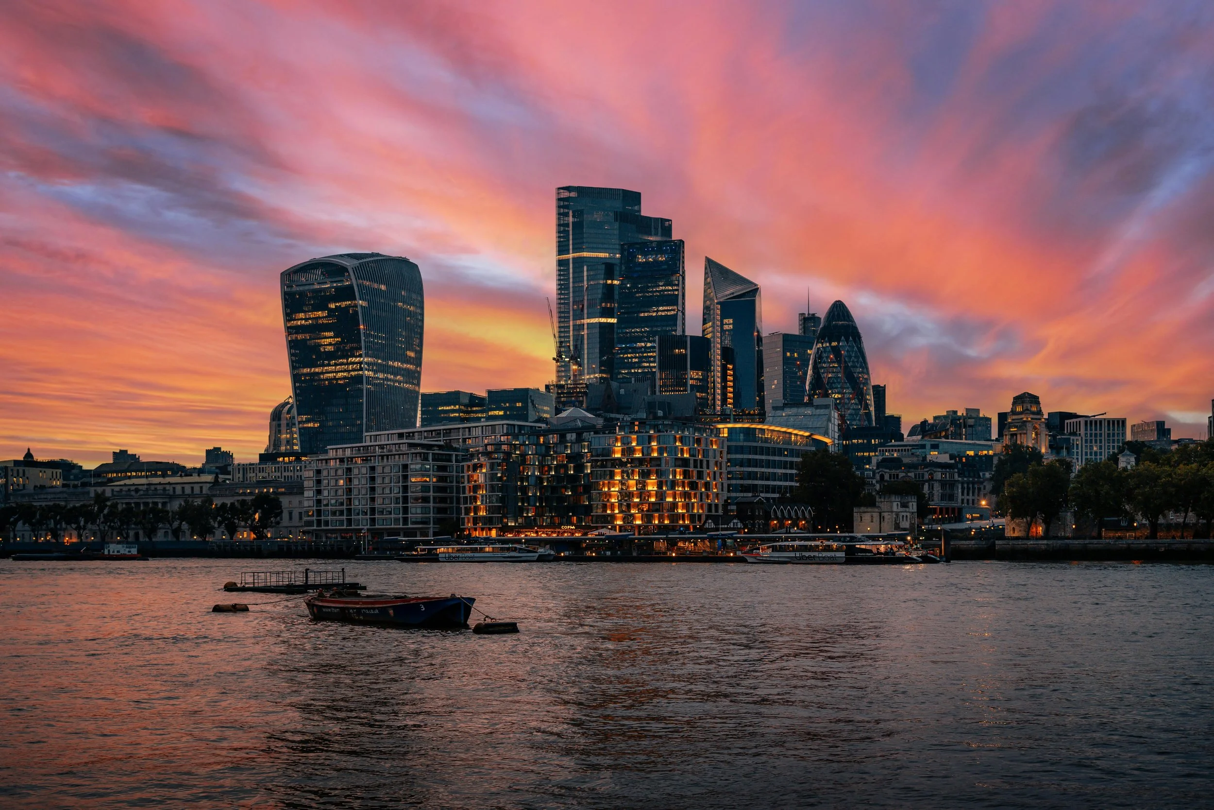 City skyline at sunset with modern skyscrapers and a river in the foreground.