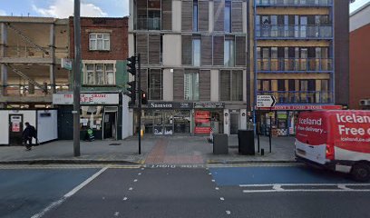 Urban street scene showing a tram stop with a large building in the background, and an iceland delivery truck on the right side.