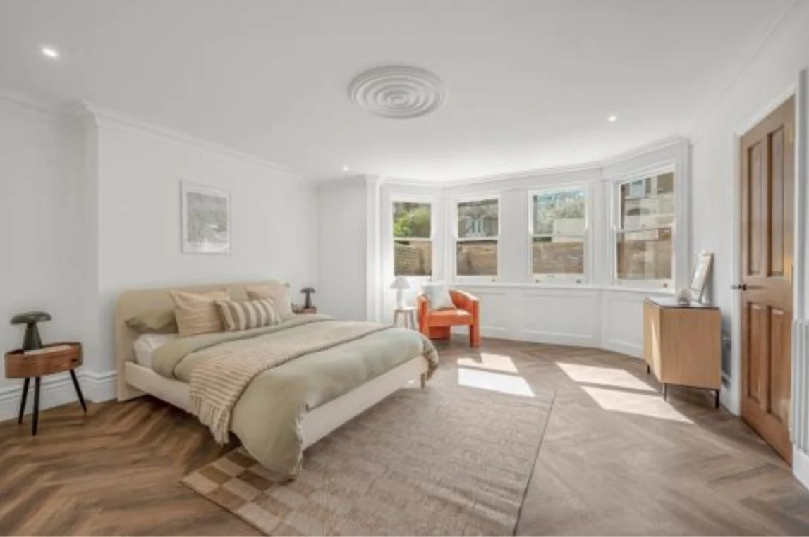 A bedroom with a bed, nightstands, a chair near a bay window, a dresser, and wood flooring.