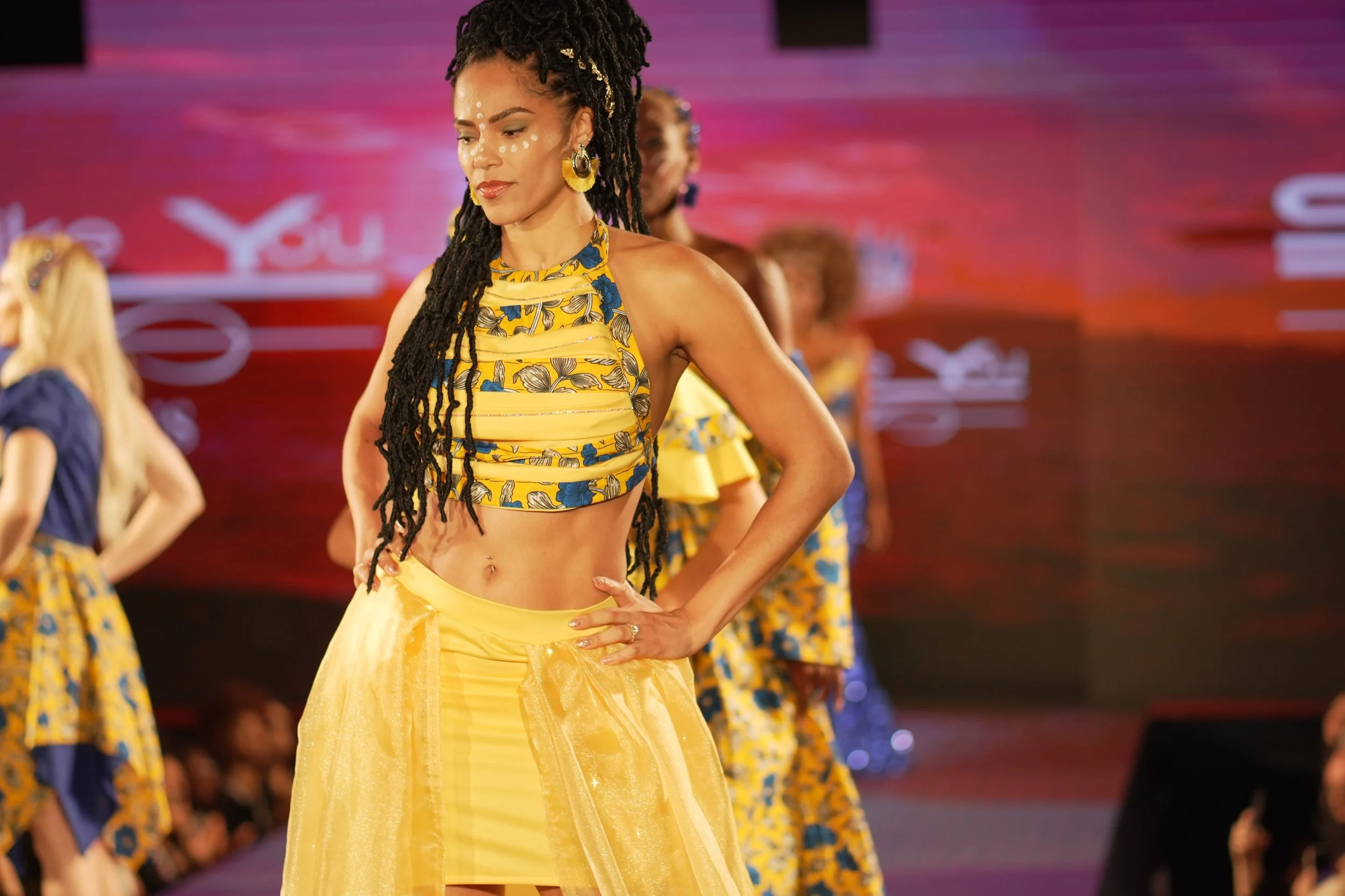 A woman with long dreadlocks and decorative face paint on her forehead is walking on a runway during a fashion show. She is wearing a yellow and blue floral crop top and a matching yellow skirt. Other models are visible in the background, also dressed in colorful outfits, with a dark purple and pink backdrop.