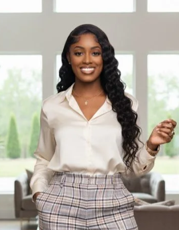 A young African American woman with long, curly black hair, wearing a cream-colored blouse and plaid pants, smiling in a bright, modern indoor space with large windows.