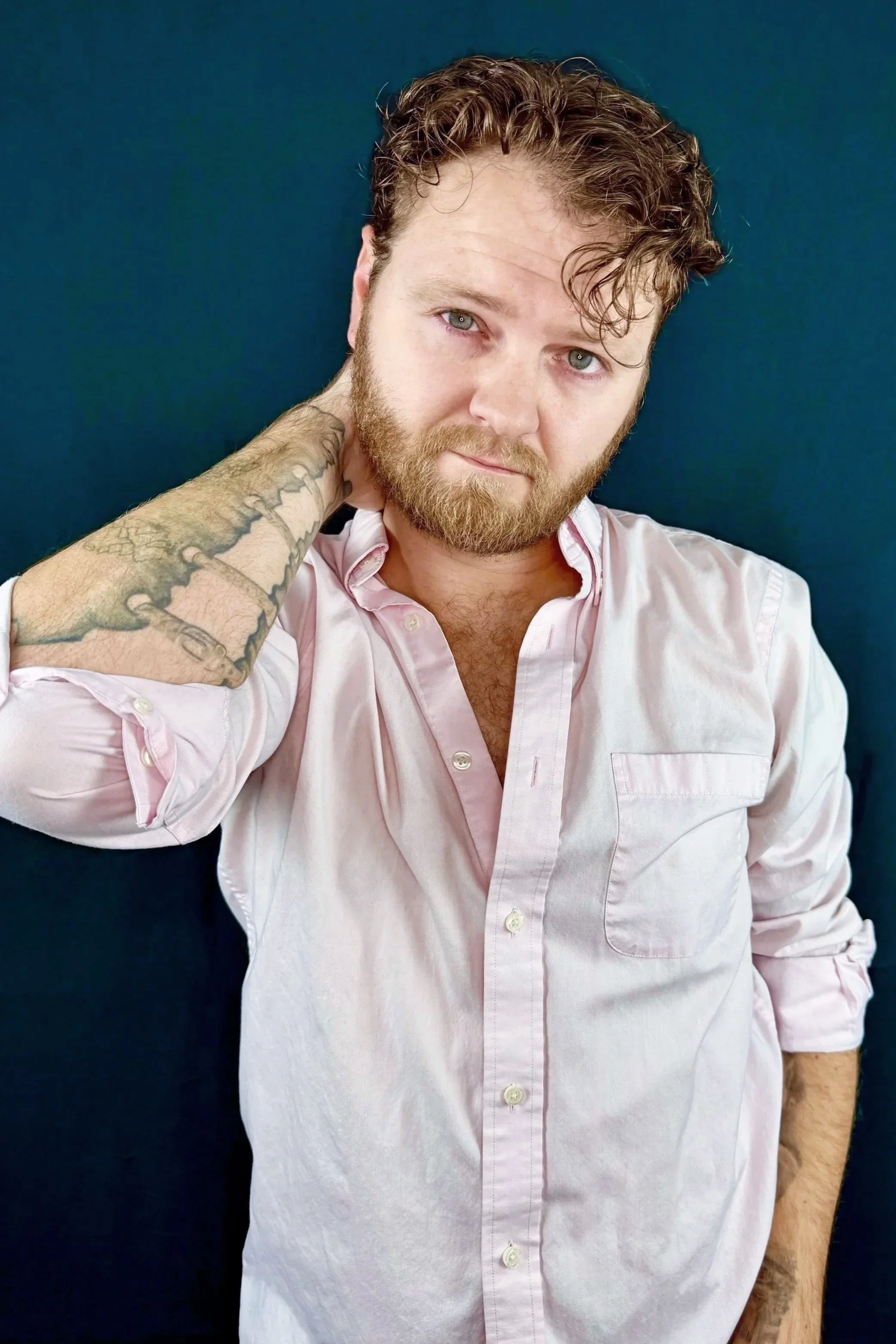 A man with curly light brown hair and a beard, wearing a light pink button-up shirt, stands against a dark blue background. He is touching the back of his neck with his right hand, revealing a tattoo on his forearm.