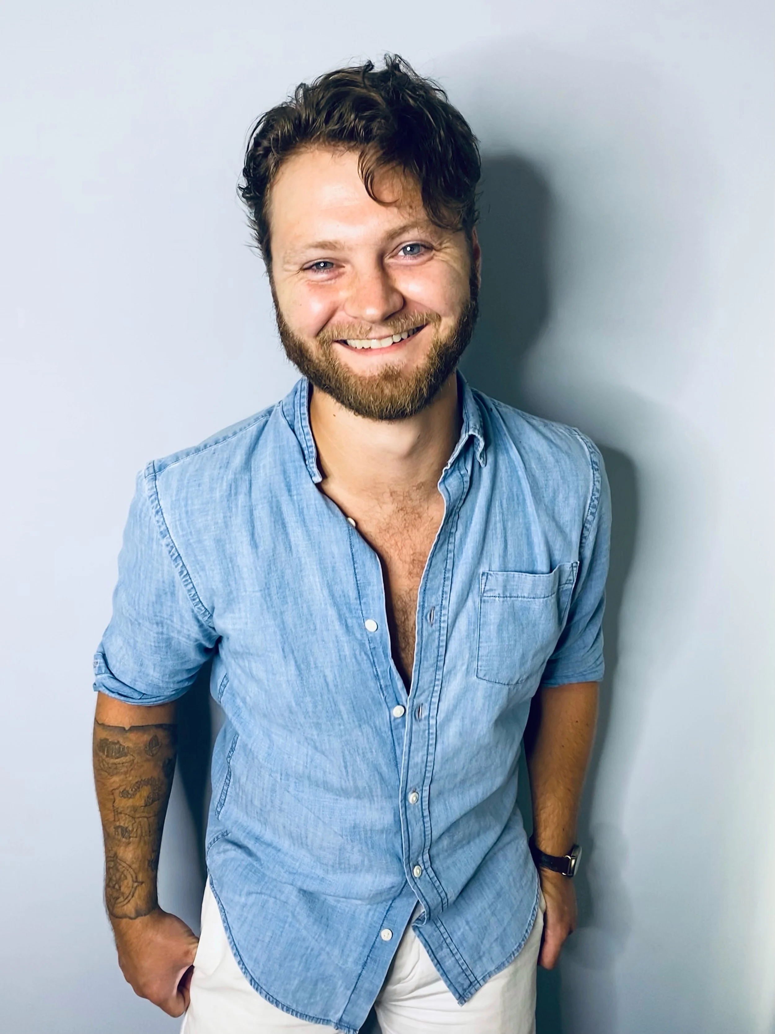 Smiling man with a beard and tattoos wearing a light blue button-up shirt and white pants, standing against a plain light-colored wall.