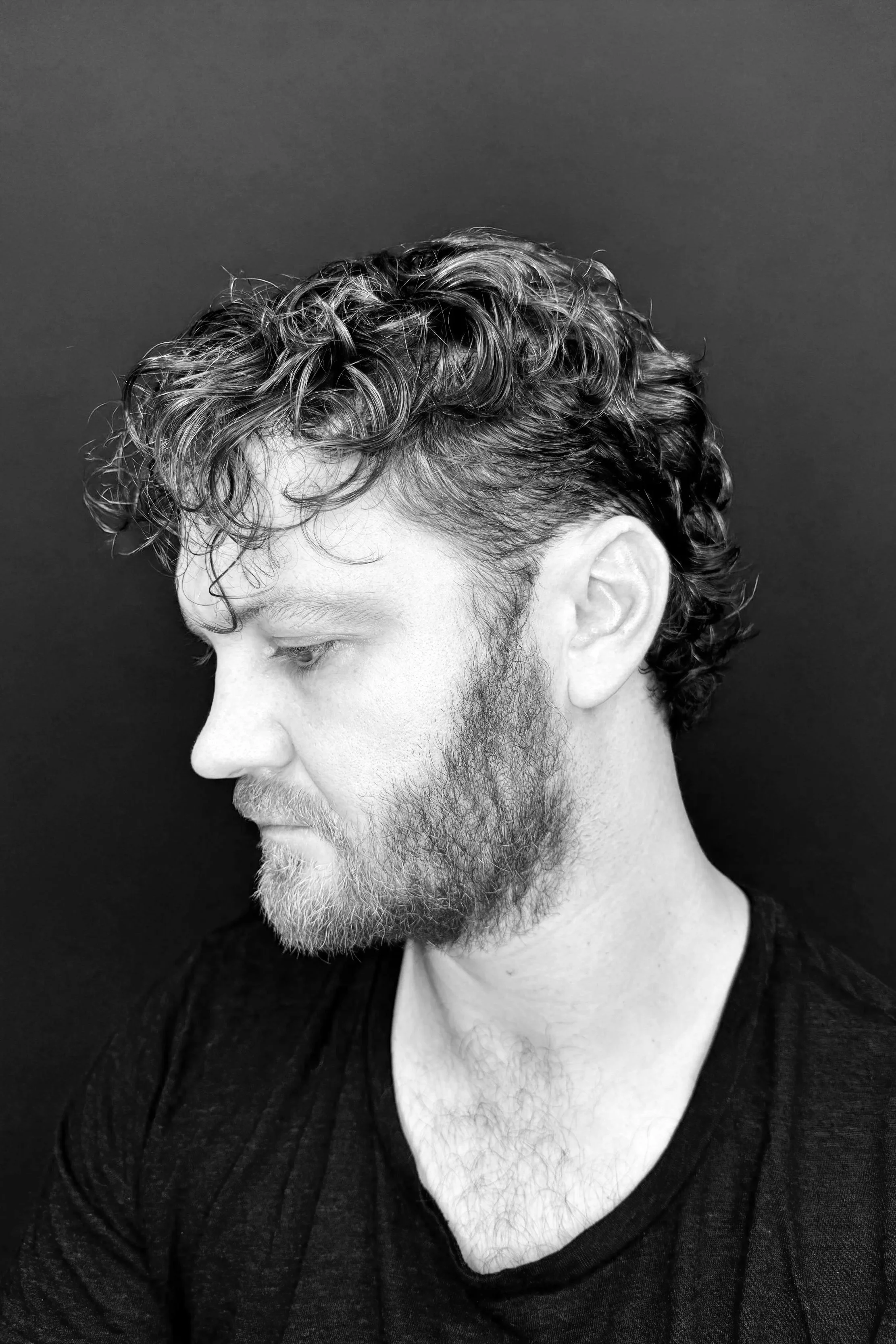A black and white profile photograph of a young man with curly hair and a beard, facing left against a plain dark background.