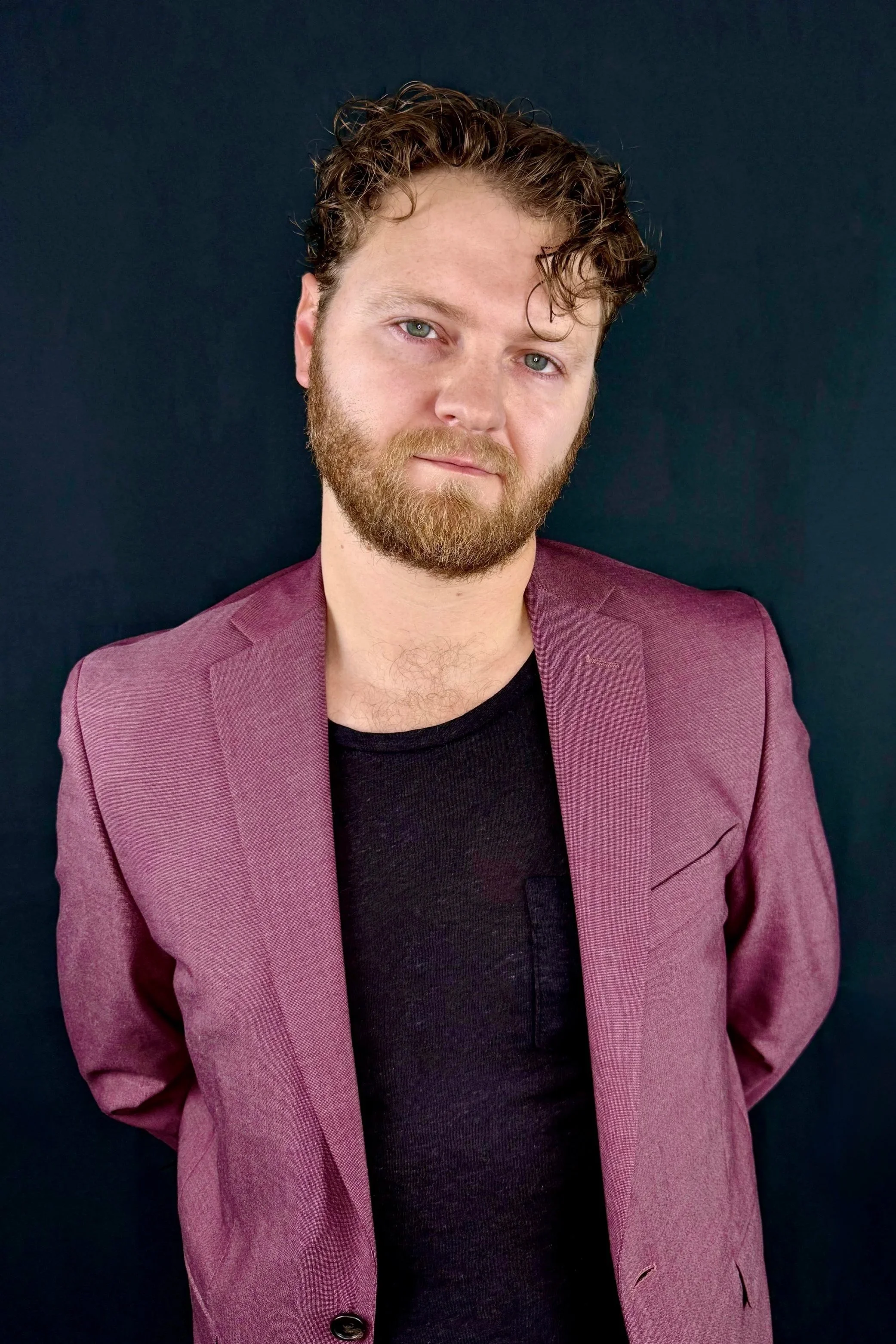 A man with curly hair and a beard wearing a maroon blazer and black t-shirt standing against a dark background.
