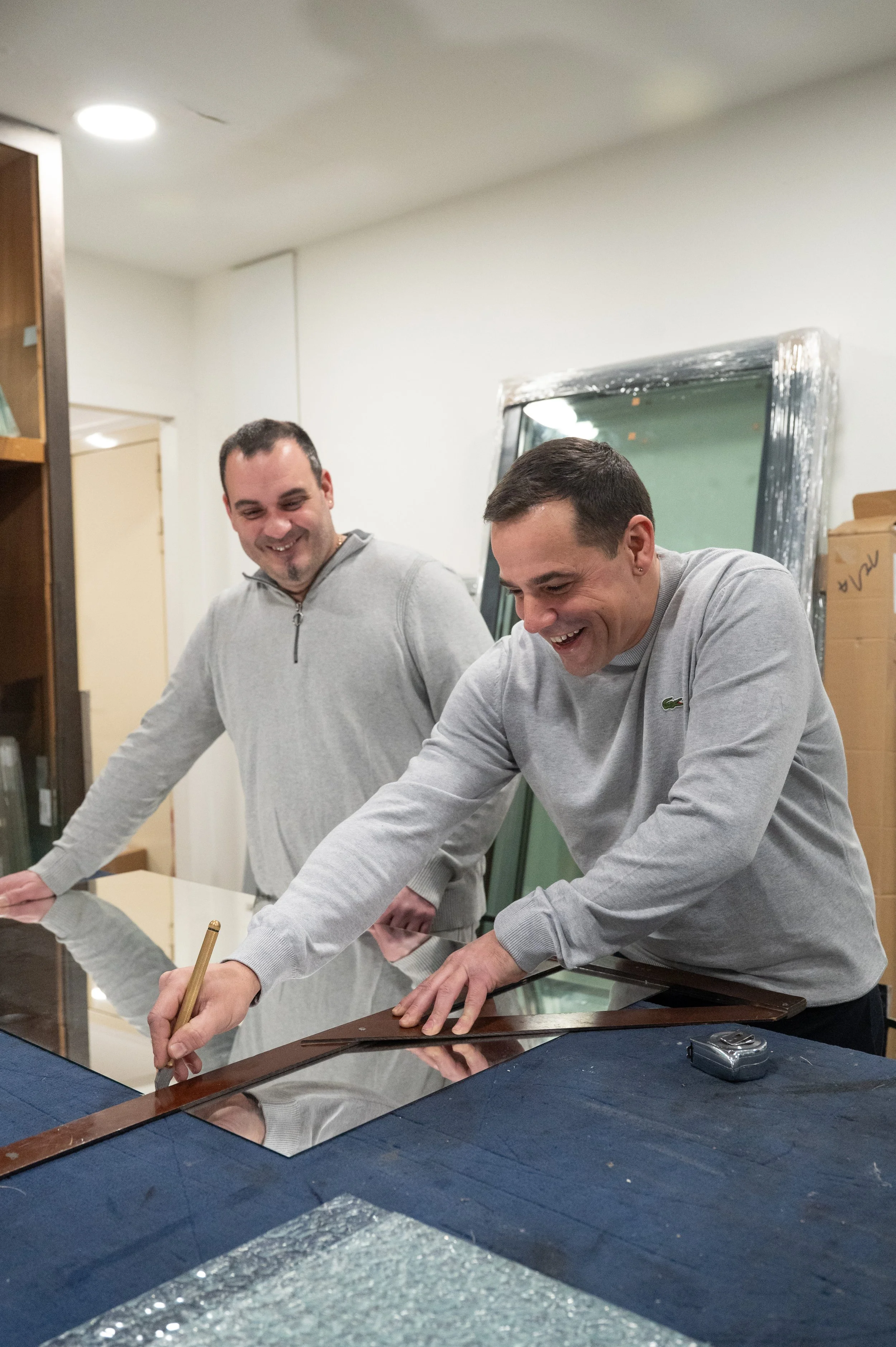 Deux hommes souriants travaillent sur une surface en verre, l'un d'eux trace avec un crayon ou un marqueur doré. La scène semble se dérouler dans un atelier ou un espace de bricolage.