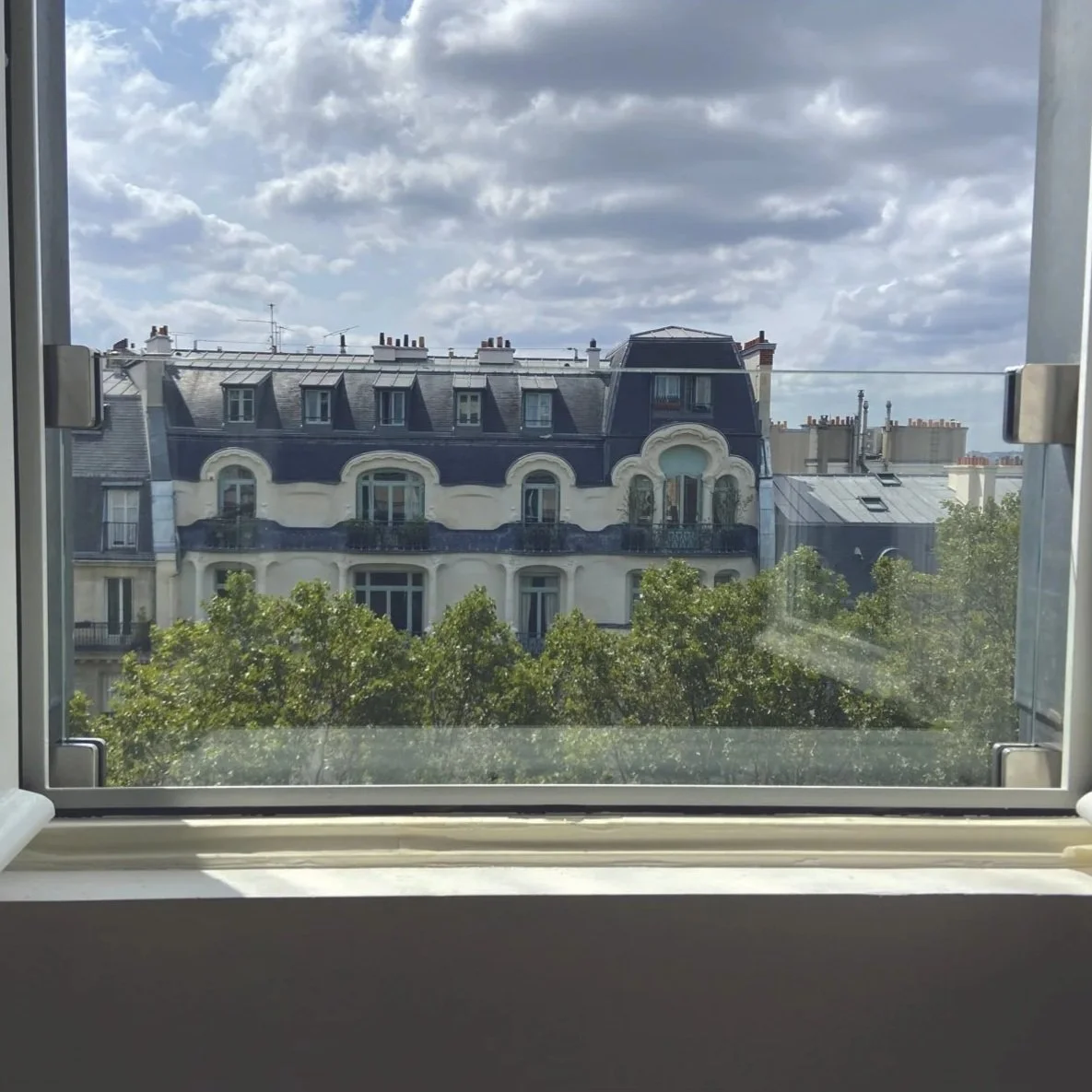 Vue d'un bâtiment haussmannien à Paris vue depuis une fenêtre ouverte, avec des nuages dans le ciel.