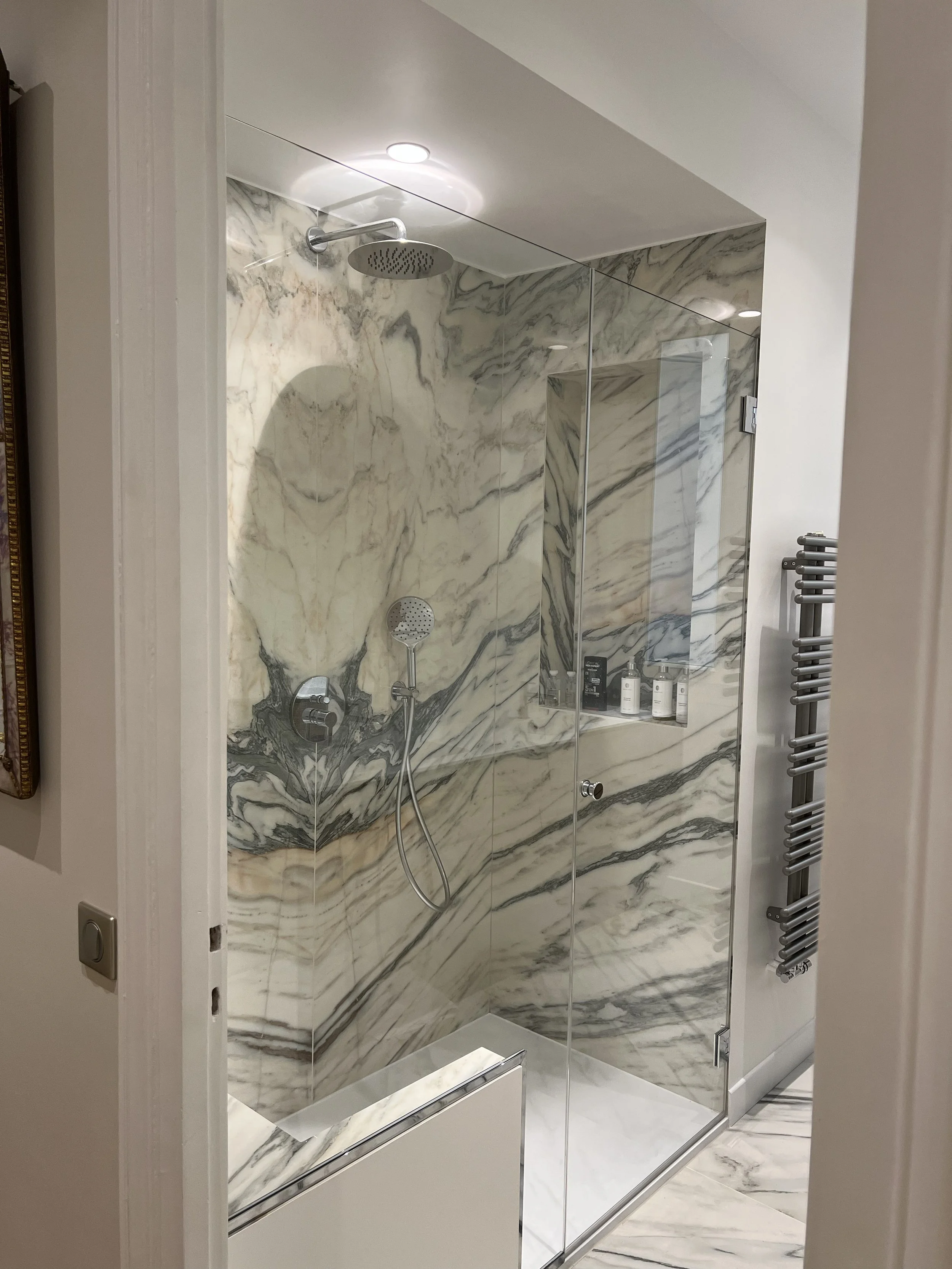 Douche en marbre avec parois en verre, plafonnier encastré, dérousse de main, étagère avec des produits de toilette, radiateur chauffant à côté.
