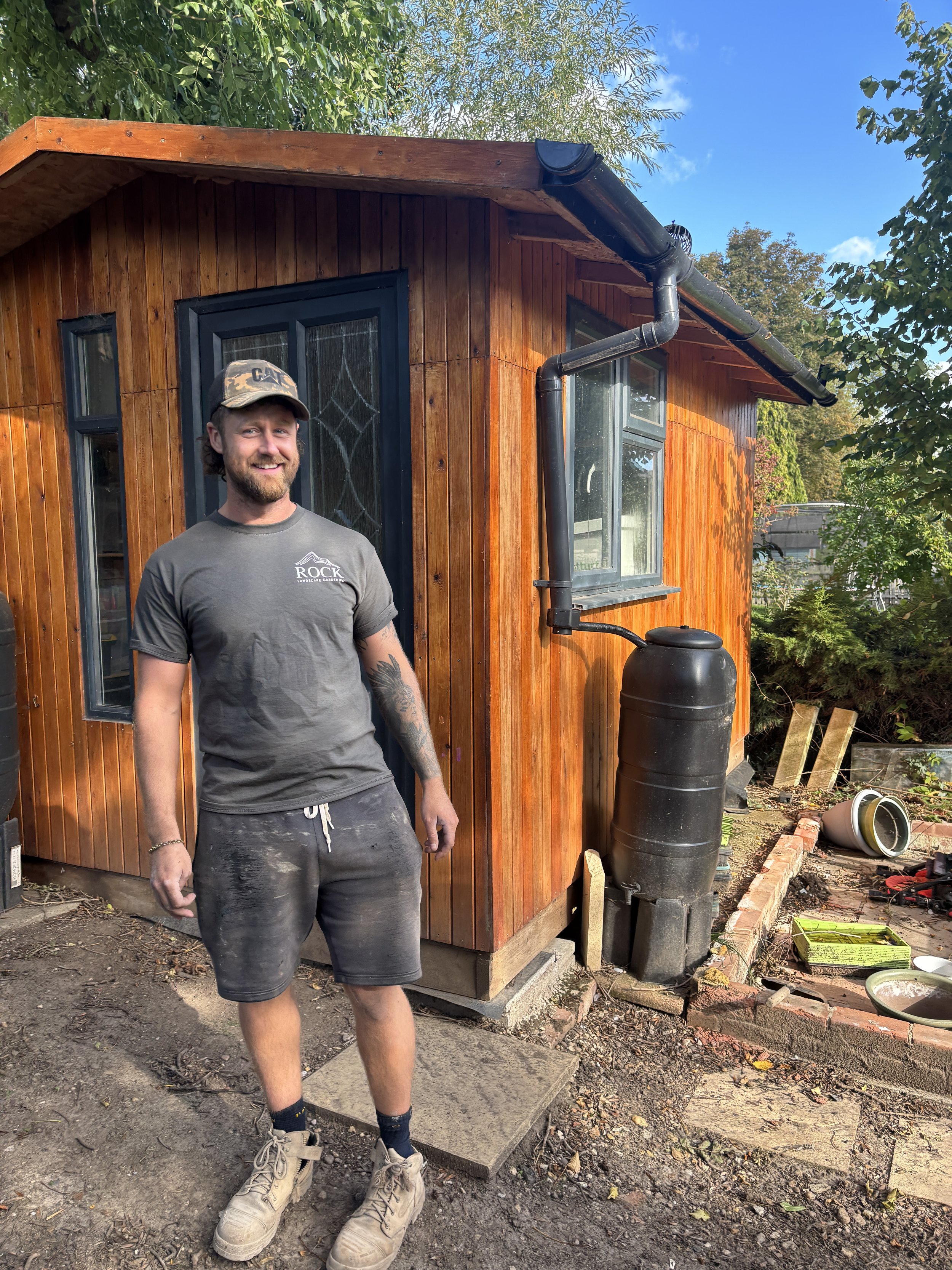 A man with a beard, wearing a gray t-shirt, shorts, work boots, and a camouflage hat, stands outside a small wooden house with a black gutter and rain barrel, smiling at the camera.