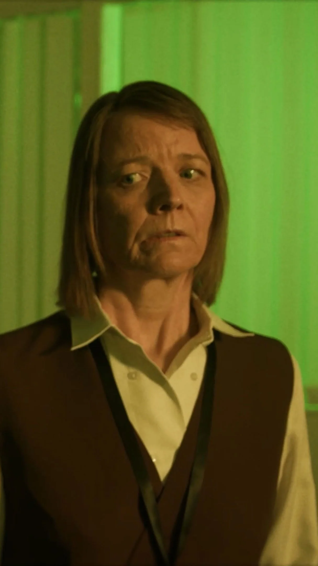 A woman with shoulder-length brown hair and green eyes stands in front of a green background, wearing a beige shirt with a dark vest.