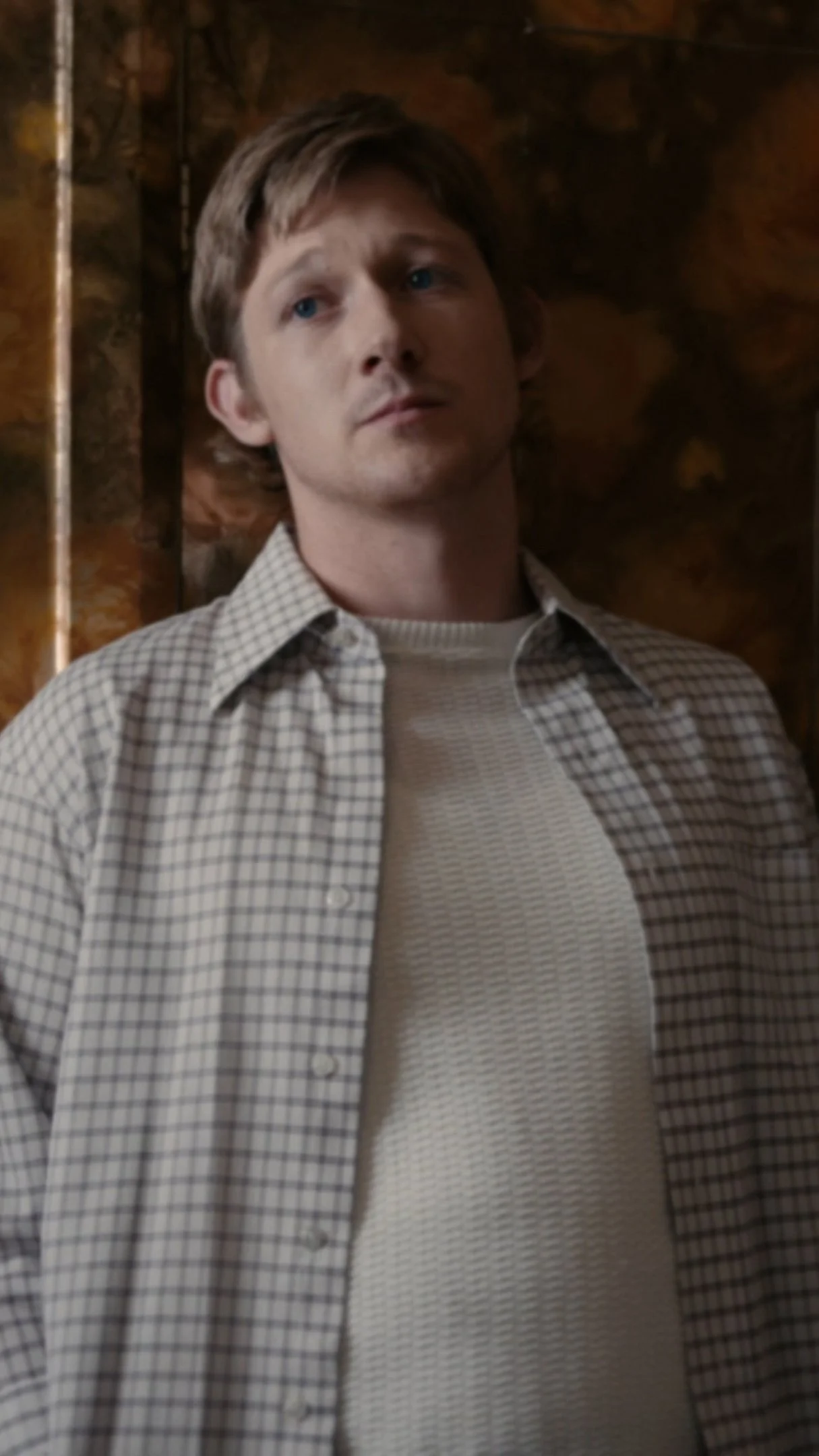 A young man with light brown hair and blue eyes wearing a plaid shirt over a textured white shirt, standing against a wooden wall.