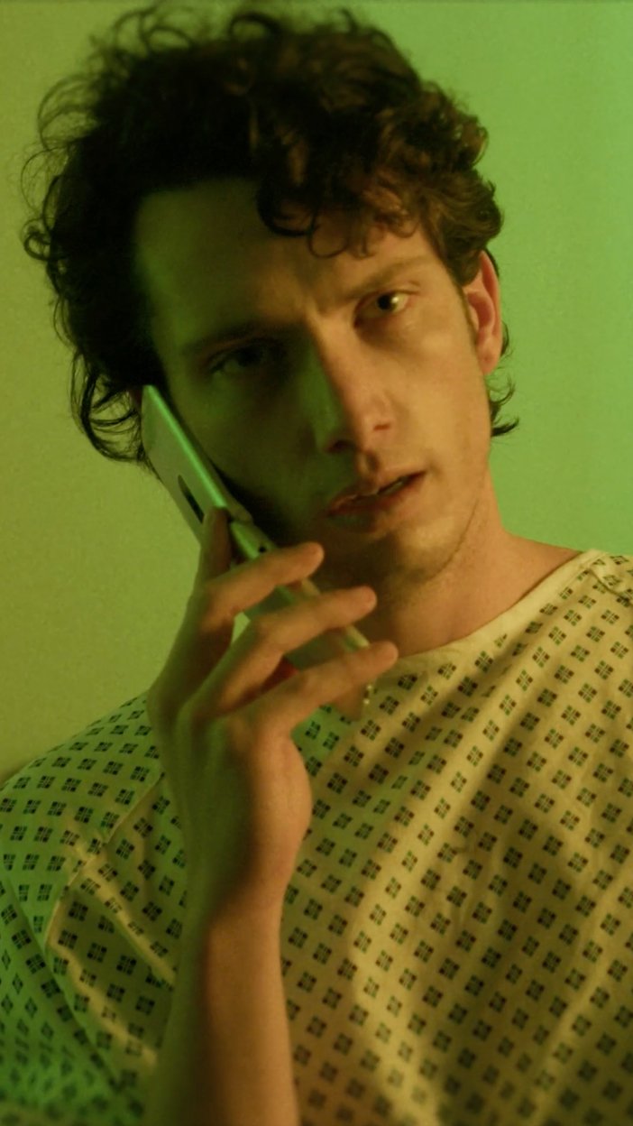 A young man with curly hair holding a phone to his ear, wearing a hospital gown against a green background.