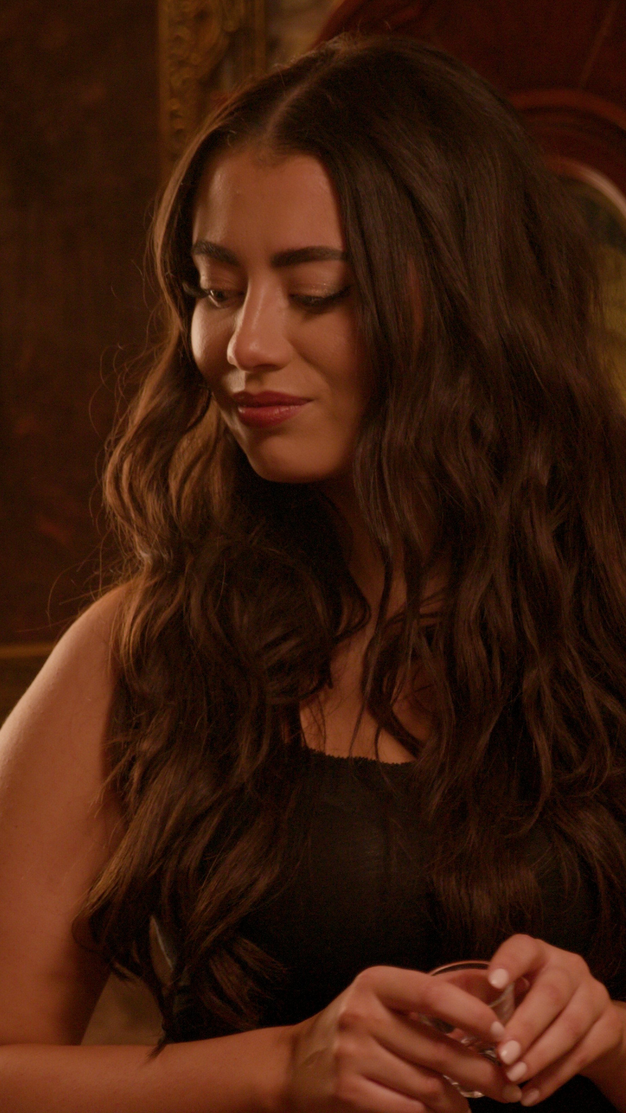 A woman with long, wavy brown hair looking down, holding a glass with both hands, in a dimly lit room.