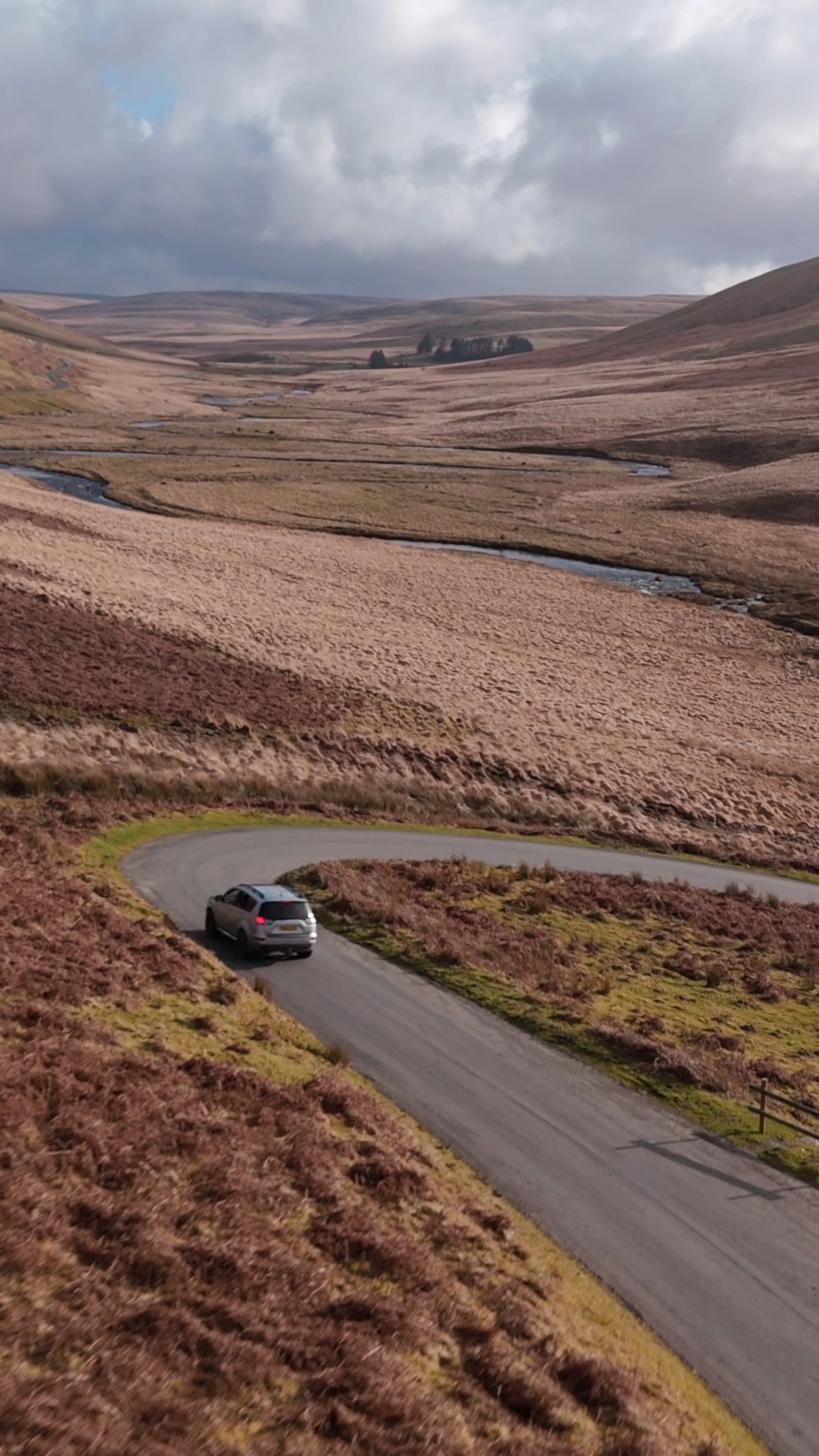 A winding mountain road with a gray SUV driving on it, surrounded by brown, grassy hills, with a cloudy sky overhead.