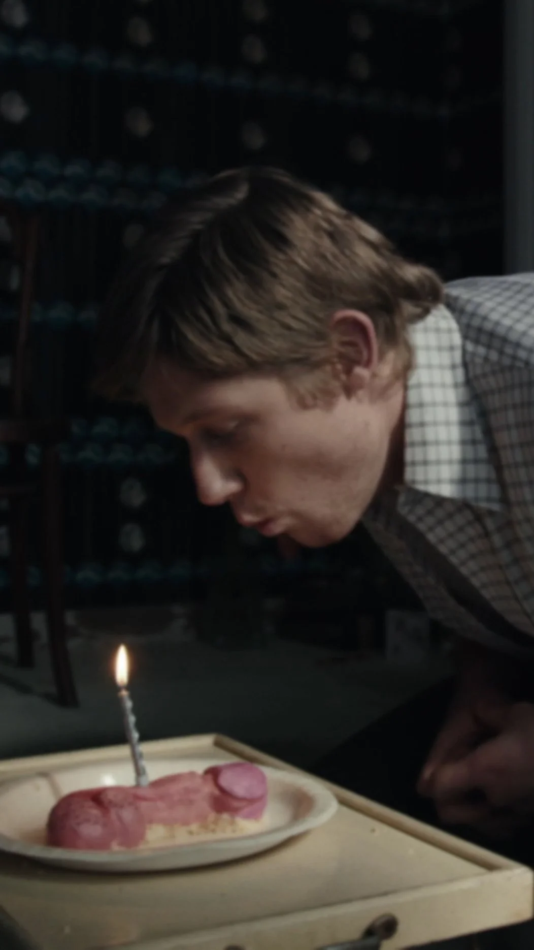 Young man leaning forward to blow out a birthday candle on a plate of food with sausage and a boiled egg, in a dimly lit room.