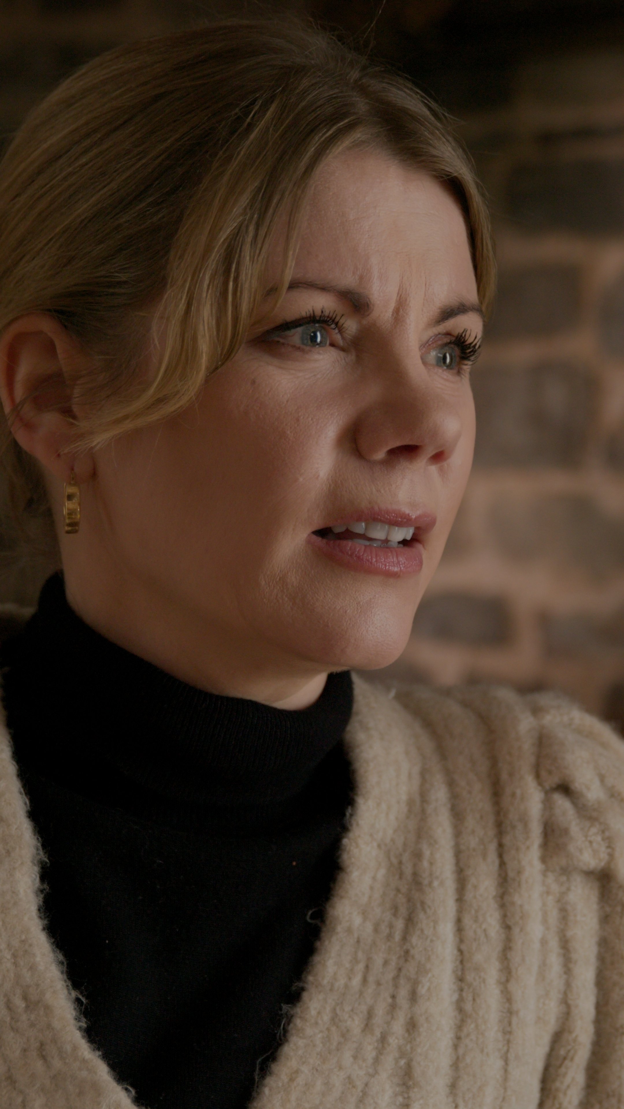 A close-up of a woman with blonde hair, wearing gold hoop earrings, a black turtleneck, and a beige fluffy cardigan, looking distressed with a blurred brick wall background.