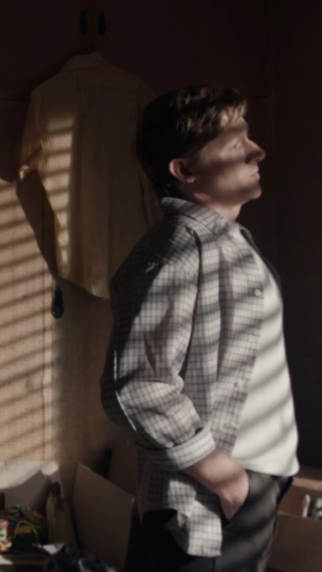 A person with short dark hair standing indoors in profile facing right, wearing a checkered shirt with the sleeves rolled up and hands in pockets. Shadows of window blinds are cast across their face and body.