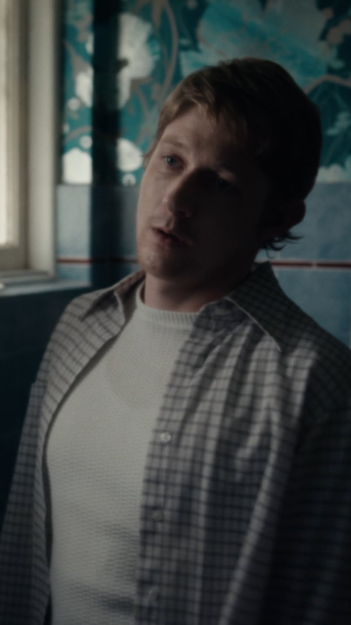 A young man with light brown hair and blue eyes looking out a window in a dimly lit room, wearing a grey and white checkered shirt over a white sweater.