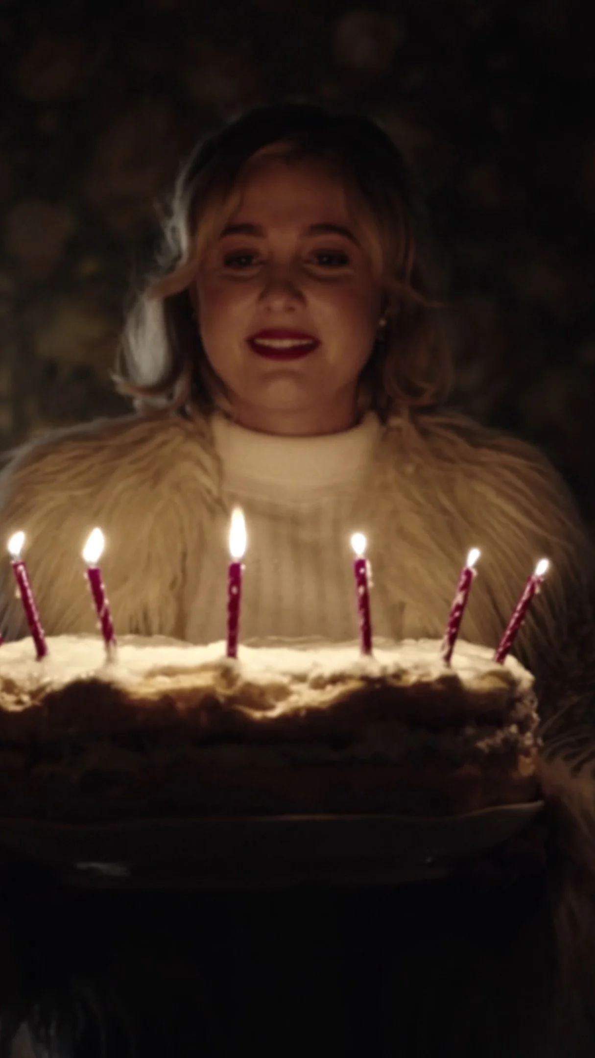 Woman with blonde hair and red lipstick holding a birthday cake with lit candles.
