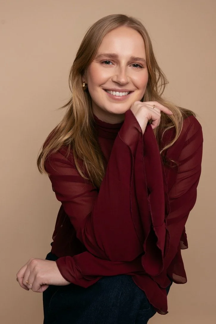 A young woman with long, wavy blond hair, wearing a burgundy sheer blouse and gold hoop earrings, smiling at the camera against a neutral background.