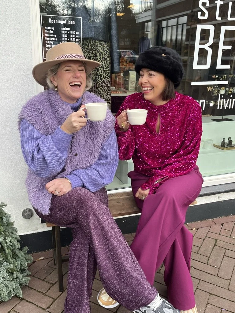 Two women sitting outside the store, smiling and enjoying coffee together. They are dressed in colorful, clothing with glitters, with one wearing a wide-brimmed hat and the other wearing a purple fur hat.