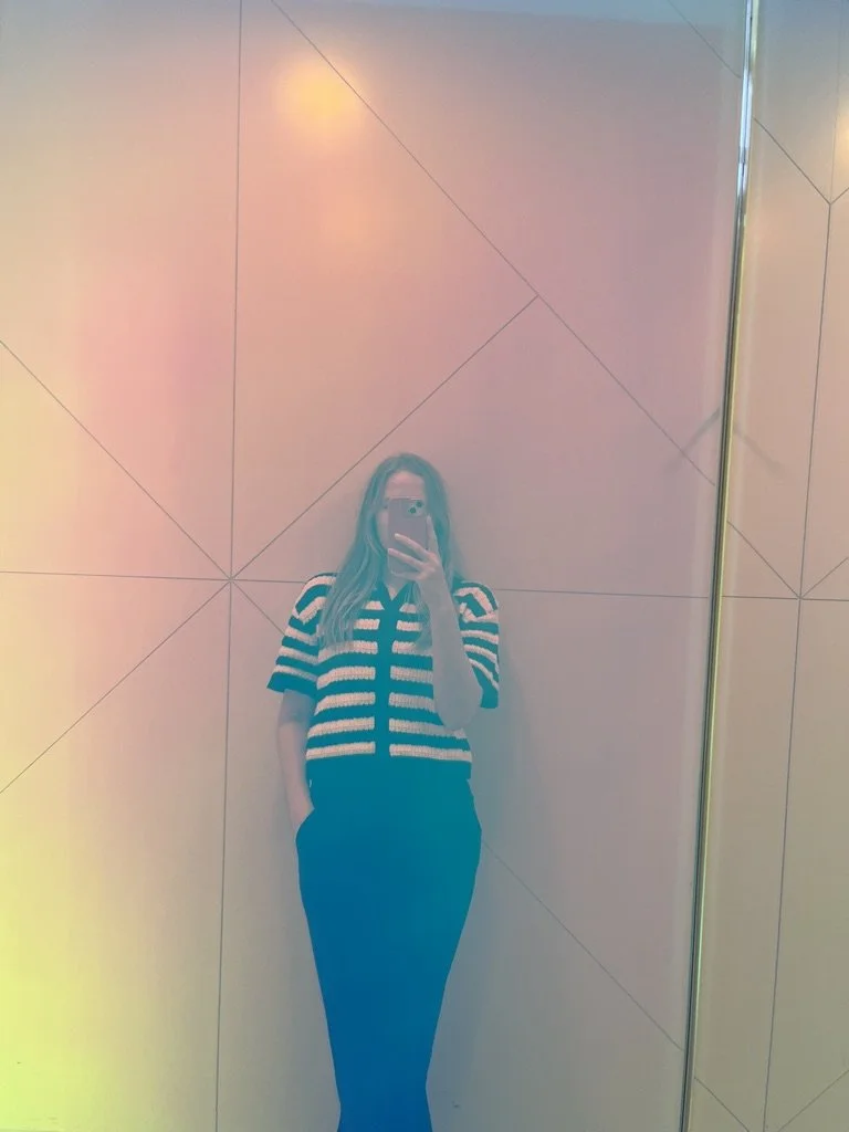 A woman taking a mirror selfie in a room with pastel-colored lighting, wearing a black and white striped shirt and black pants.