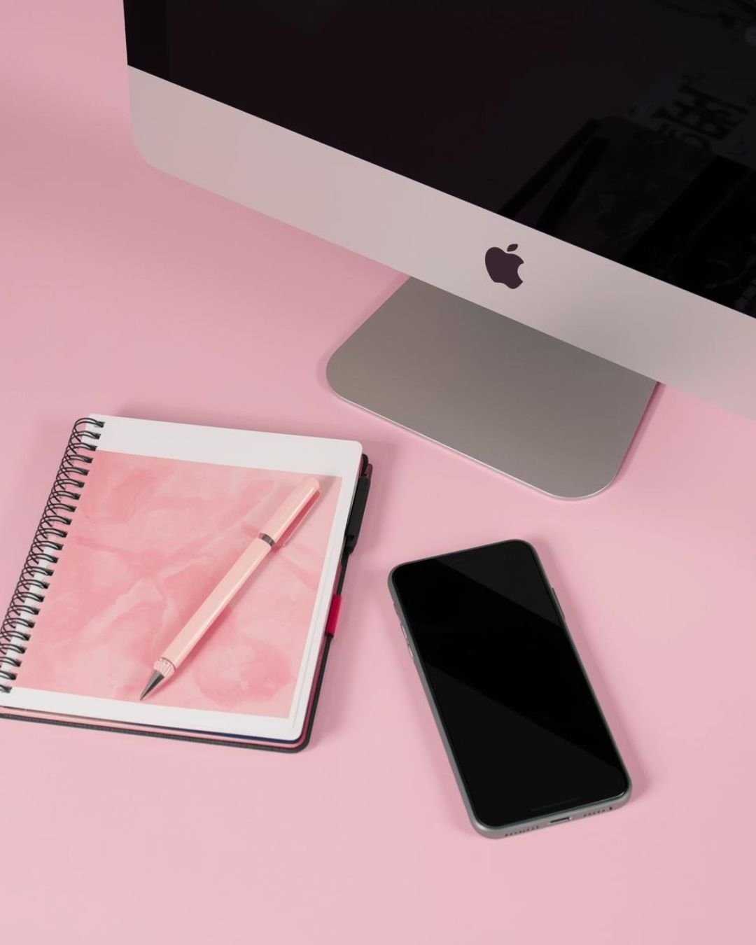 Computer monitor, smartphone, pink notebook with pen, and black pen on a pink desk.