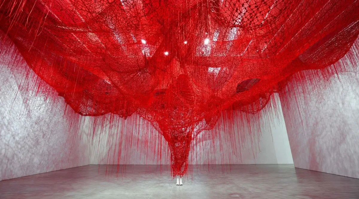 Large red art installation made of intersecting lines and strings hanging from the ceiling, creating abstract shape in a gallery with white walls and hardwood floor.