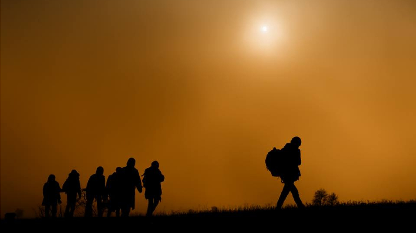 Silhouettes of six immigrants walking on a hill during sunset with an orange sky and the sun visible.