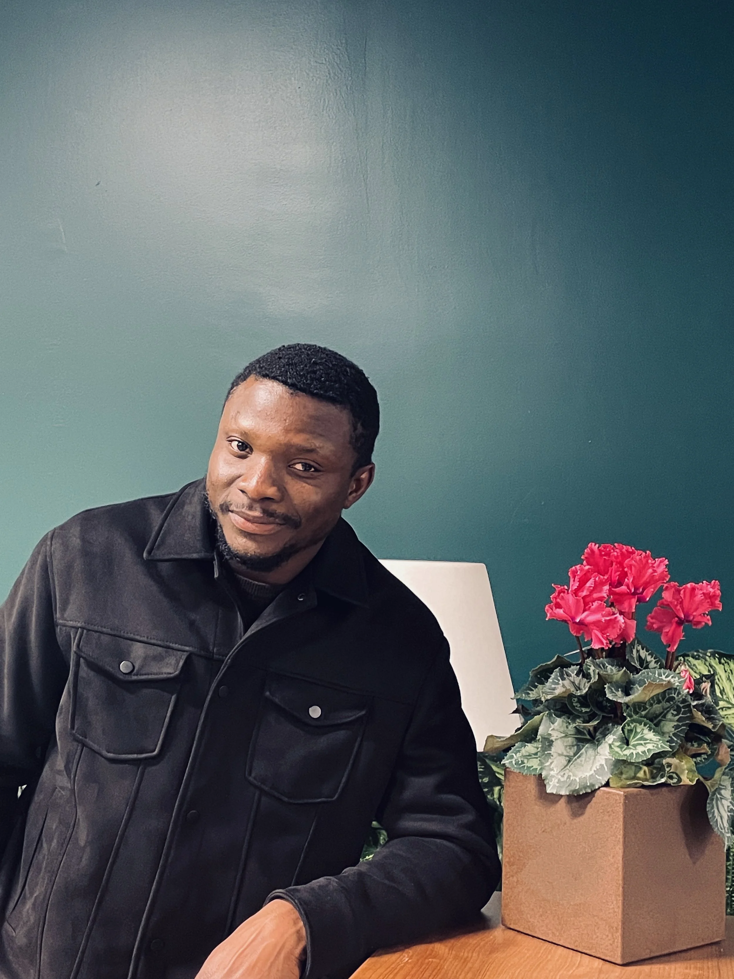 A man leaning on a wooden surface next to a potted pink cyclamen plant, against a green wall.