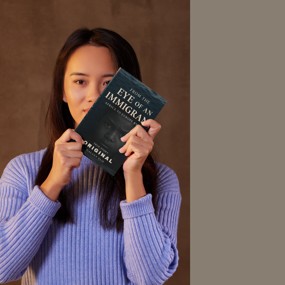 A woman with long dark hair holding a book titled "From the Eye of an Immigrant" in front of her face, revealing only her eyes and part of her forehead. She is wearing a light purple ribbed sweater and stands against a brown background.