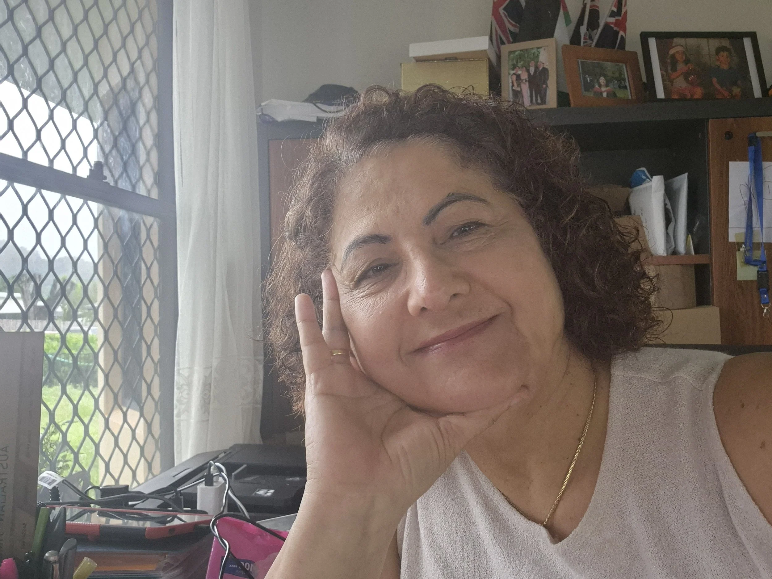 A relaxed portrait of a woman with curly hair resting her face on her hand, seated in a softly lit interior space that conveys warmth, reflection, and personal presence.