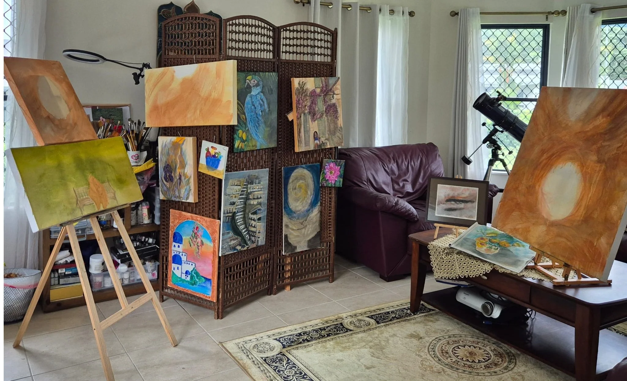 A room with several paintings, some displayed on easels and a folding screen. There is a wooden table and a leather couch, with a telescope near the window. Natural light filters through white curtains.