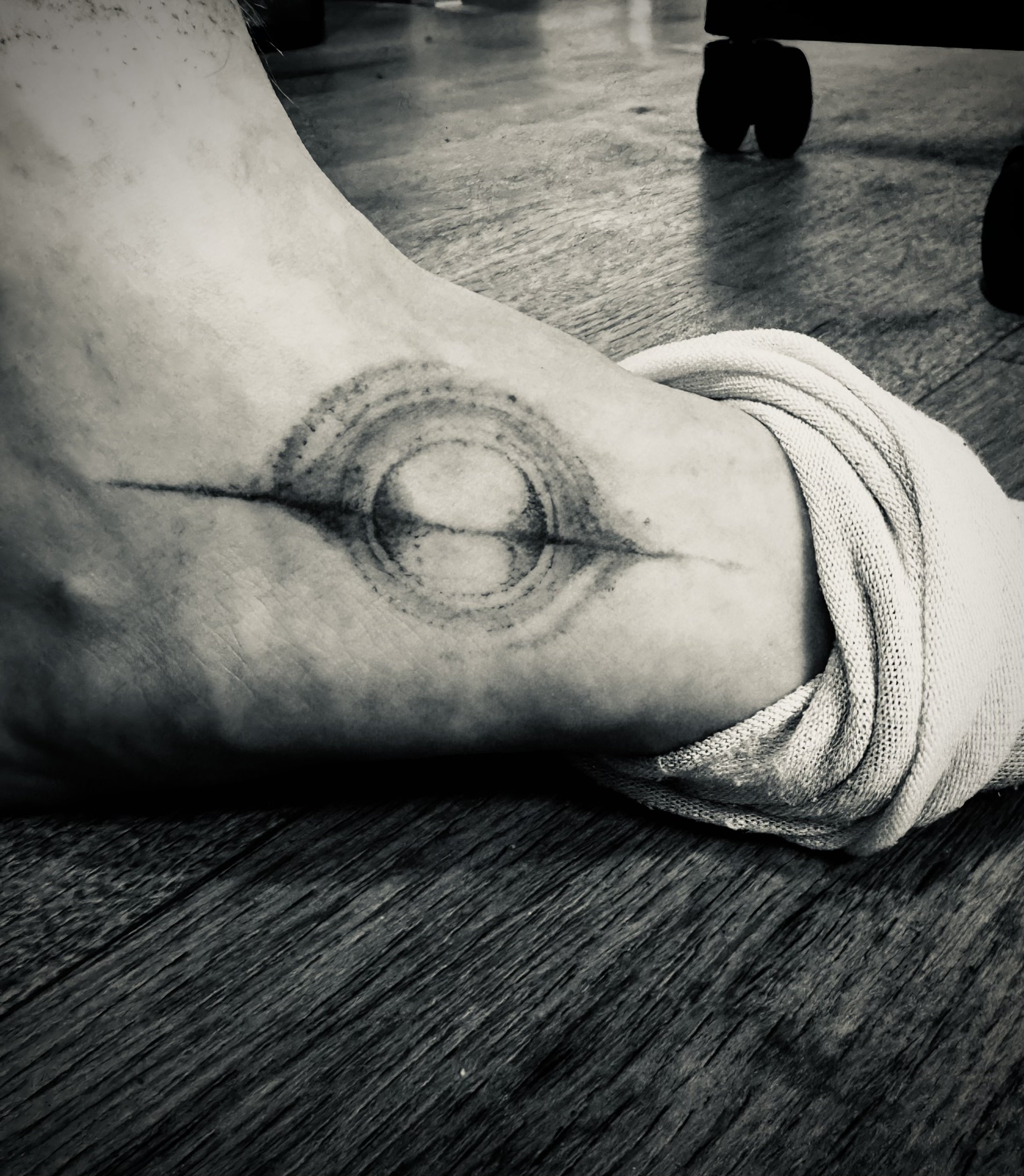 Schwarz-weiß Foto eines Handgelenks mit einem Tattoo eines digitalen Kreis-Designs, auf einem Holzfußboden liegend, sichtbar sind auch die Pantoffeln im Hintergrund.