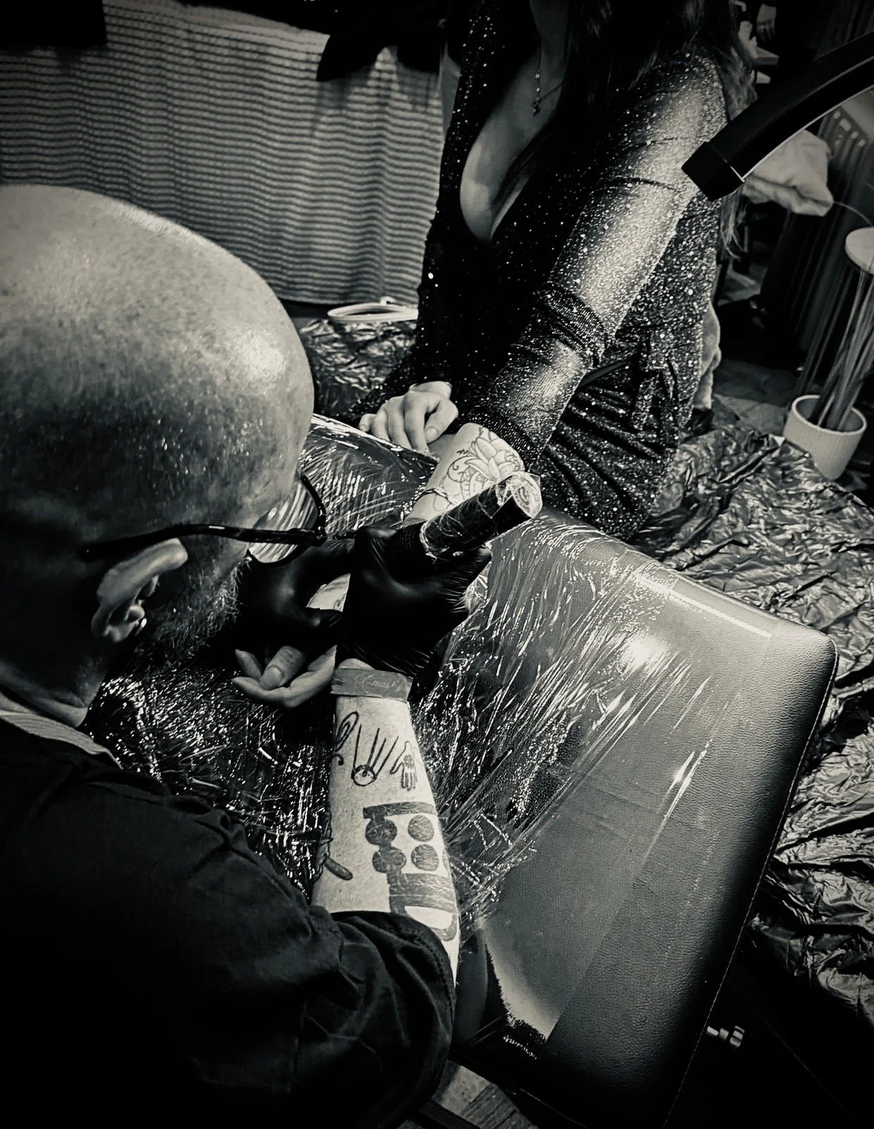Ein Tätowierer mit Glatze, Brille und Tattoos auf den Armen, tätowiert einen Kunden am Arm. Der Kunde trägt ein dunkles, glitzerndes Outfit und sitzt auf einem Stuhl, während der Tätowierer mit einem Tattoo-Maschinenarm arbeitet. Die Szene ist in Schwarz-Weiß aufgenommen.