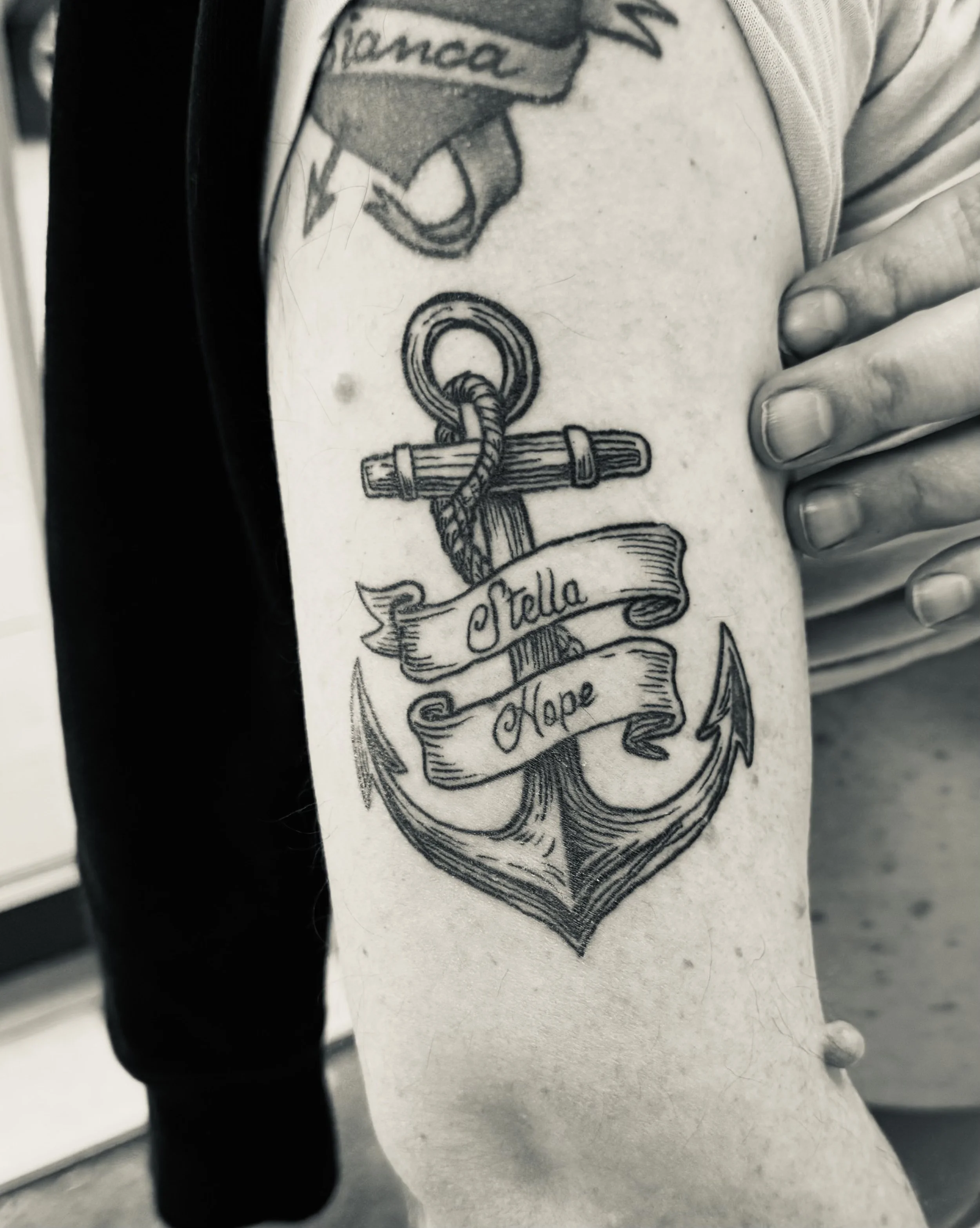 Schwarze Tätowierung eines Ankers mit einer Schleife, auf der die Worte 'Stella' und 'Hope' geschrieben sind, auf dem Oberarm.