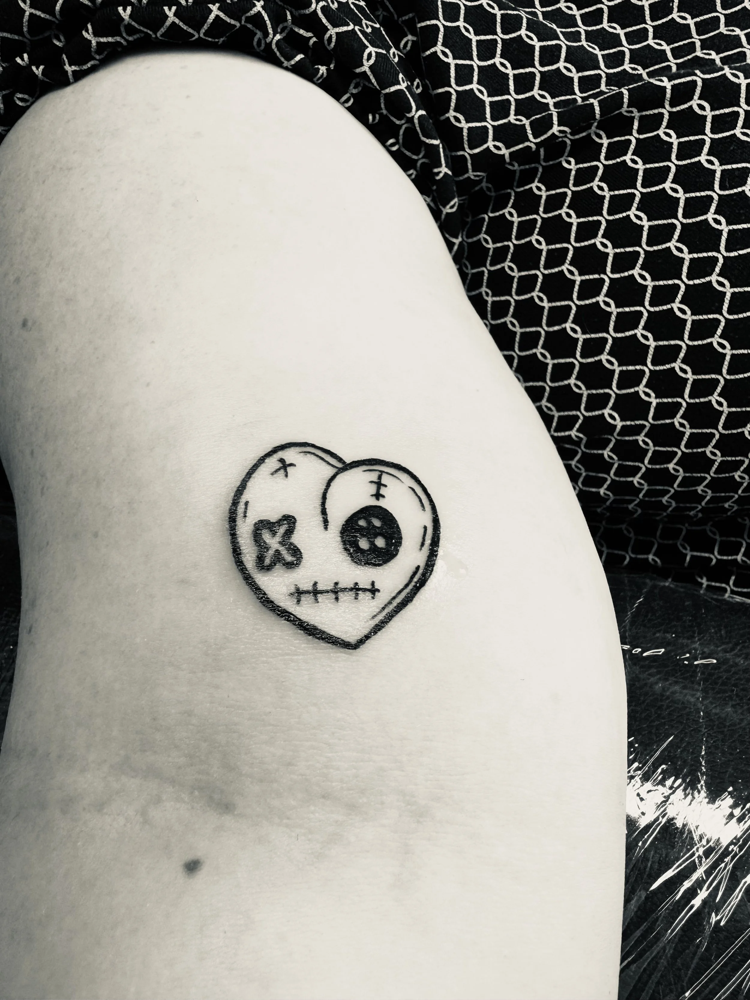 Schwarzes Tattoo in Form eines Herzens mit einem Gesicht, das halb Gesichtsausdruck zeigt, auf einer Person mit heller Haut, umgeben von schwarzem Stoff mit weißen Mustern.